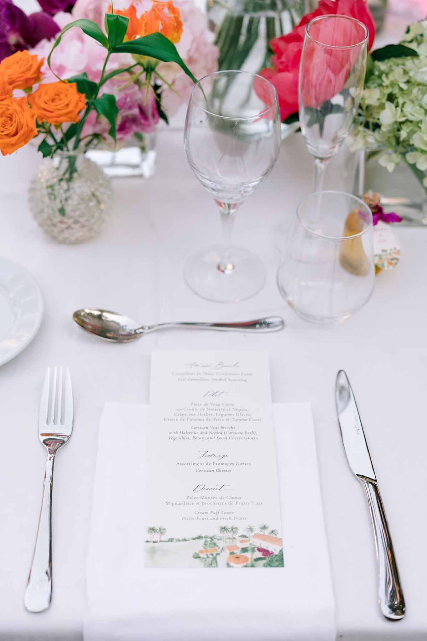 Reception place setting with French menu card, silver flatware, and bold orange and pink floral arrangements