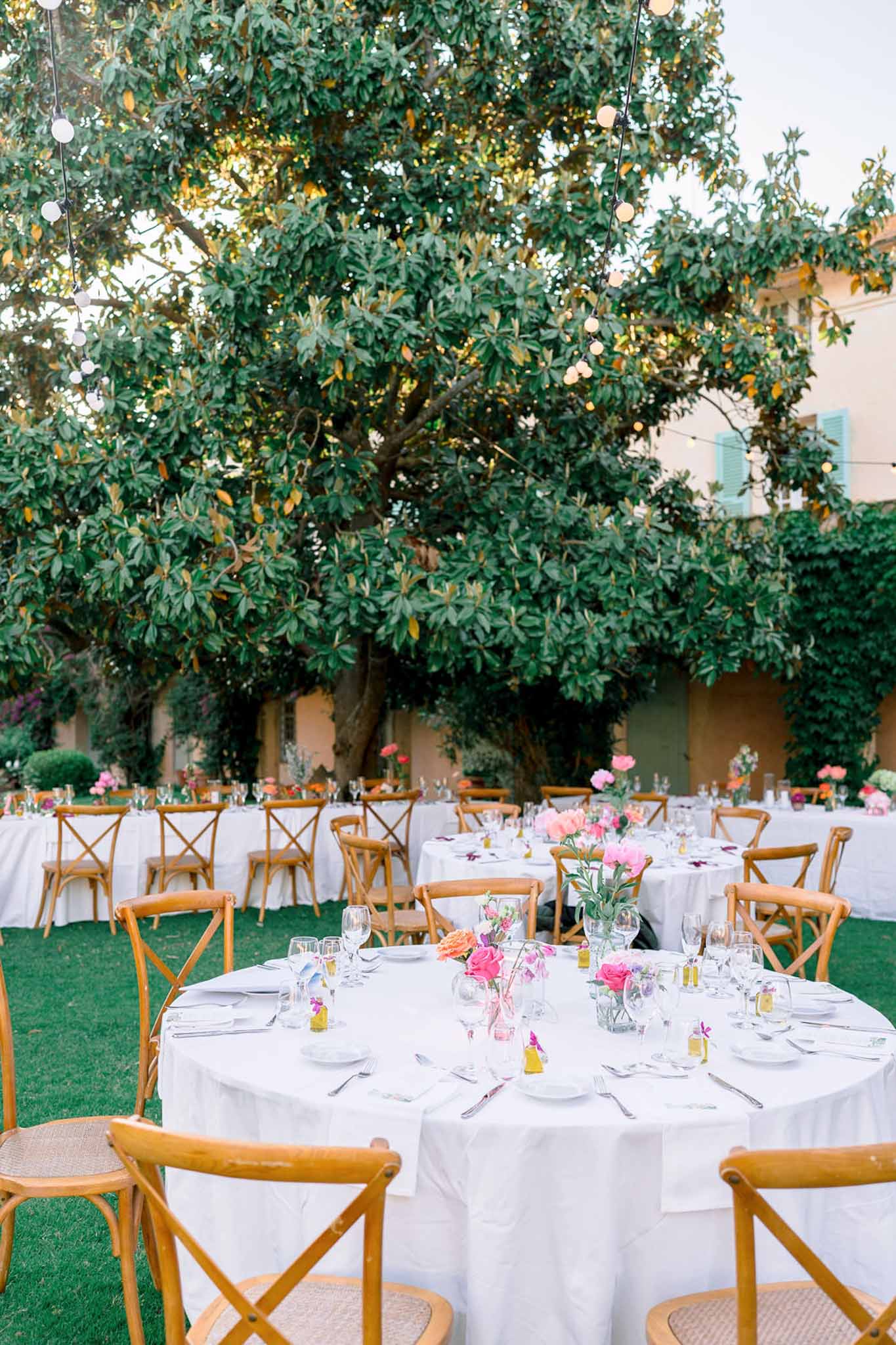 Round reception tables with cross-back chairs and pink peony centerpieces in a manor courtyard with string lights