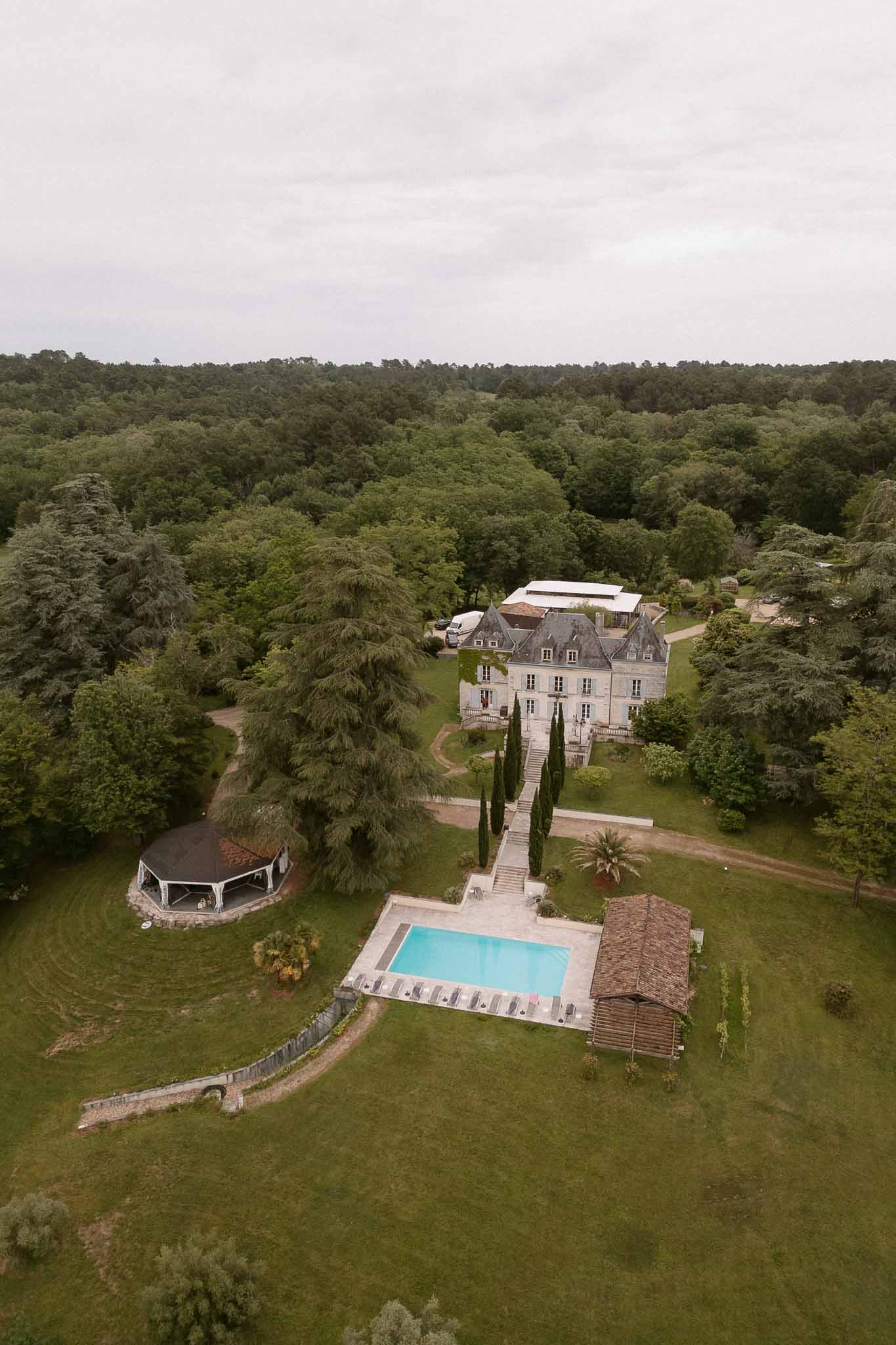 Aerial chateau with slate turrets, pool terrace, octagonal gazebo, and white event marquee in woodland