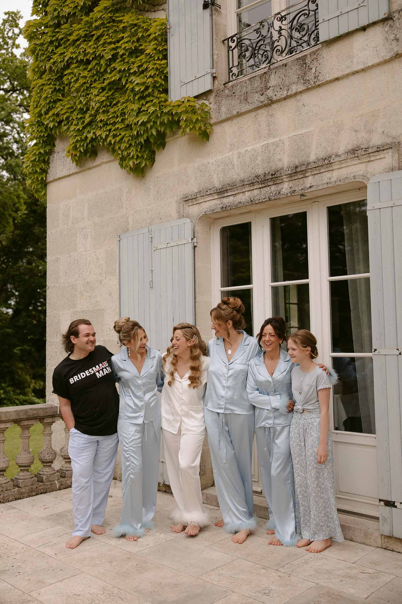 Bride and five bridesmaids laughing in matching satin pajamas on chateau stone terrace