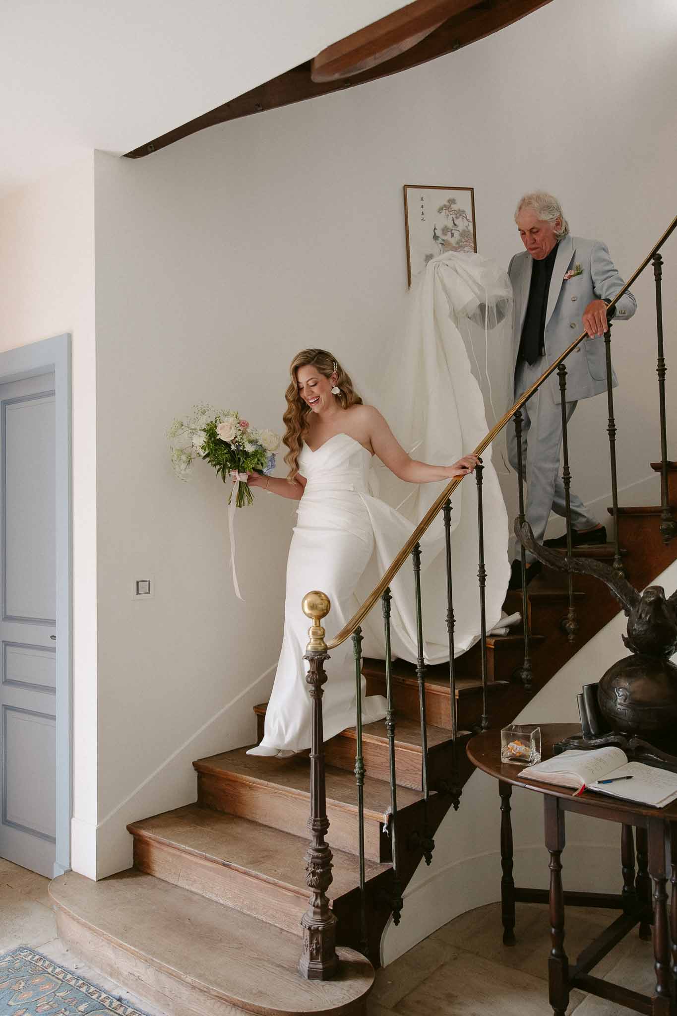 Bride laughing while descending curved wooden staircase in strapless satin gown as man carries her train behind her