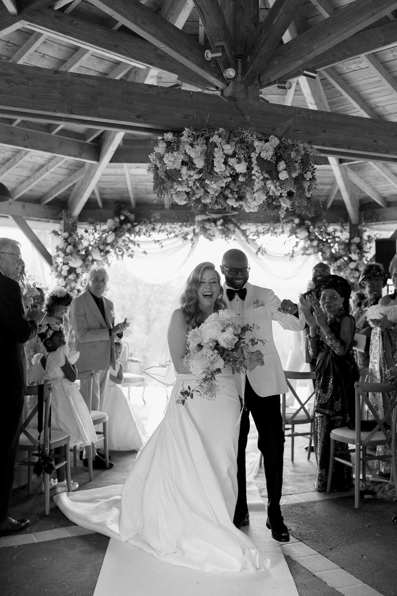 Black and white recessional walk under wooden pavilion with suspended floral chandelier and applauding guests
