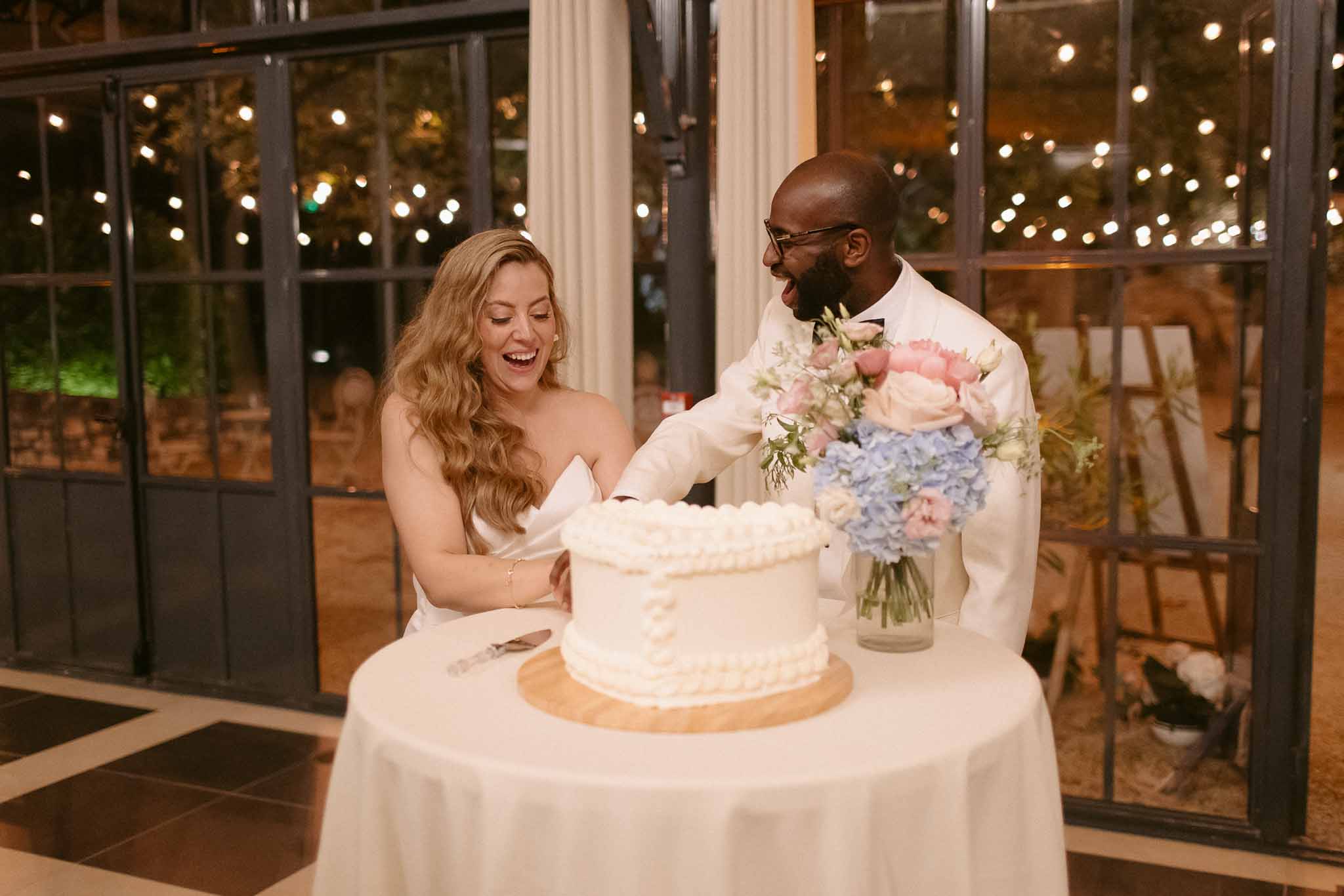 Bride and groom laugh while cutting single-tier white wedding cake at indoor evening reception