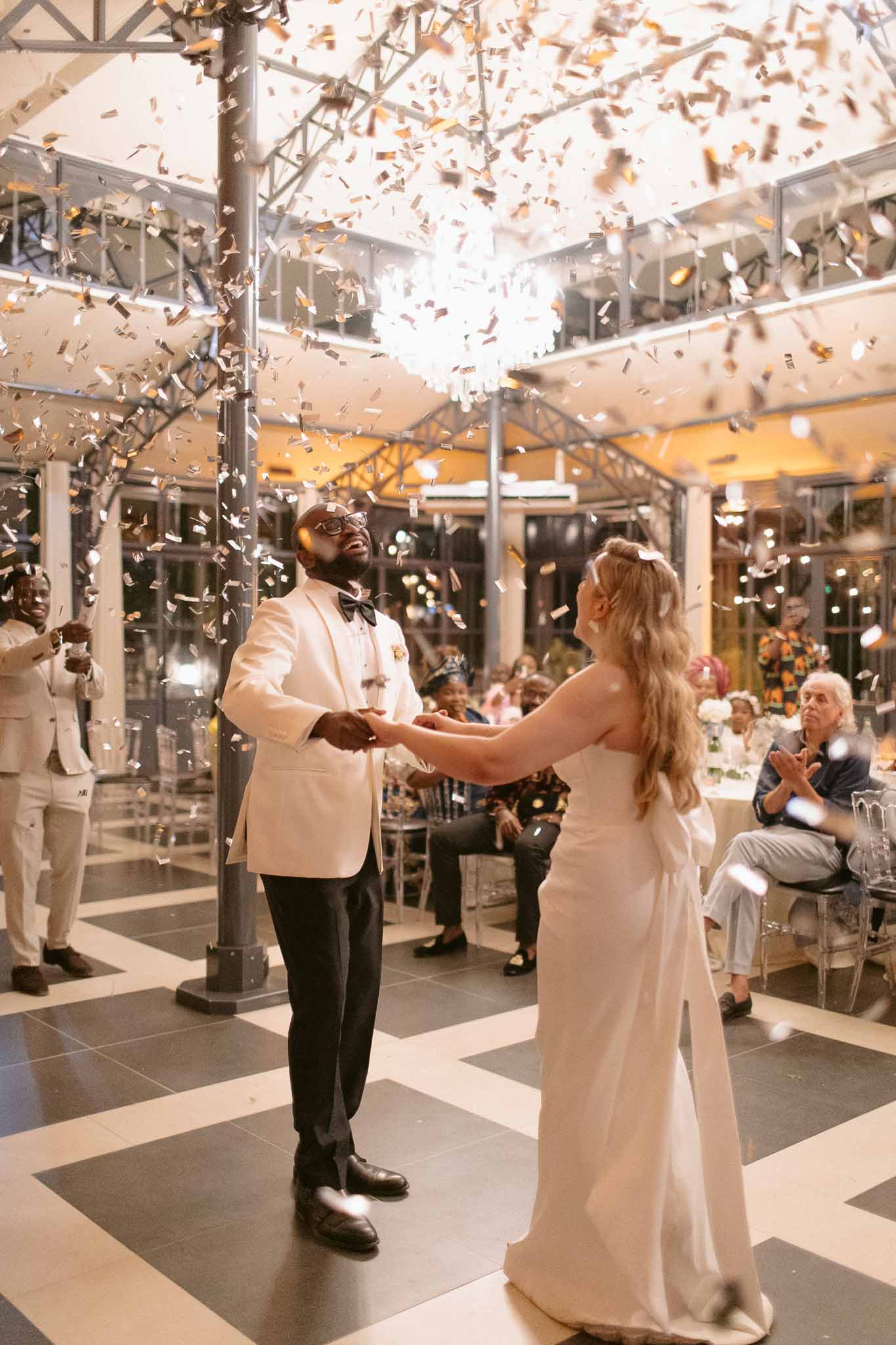 Gold confetti shower during first dance in glass-roofed venue with chevron floor and ghost chairs