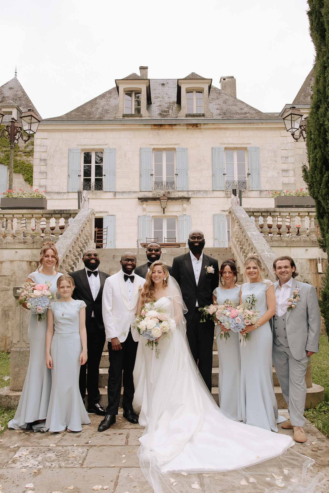 Bridal party of nine posed at base of chateau stone staircase with bridesmaids in dusty blue gowns