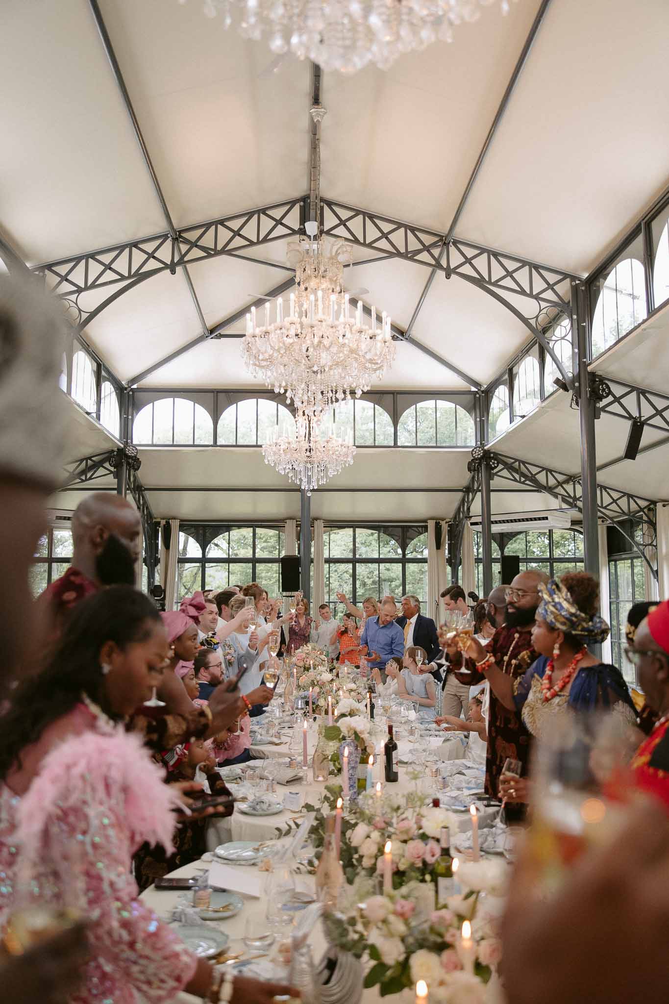 Guests raise glasses in a toast at a long banquet table inside a greenhouse venue with crystal chandeliers and blush florals