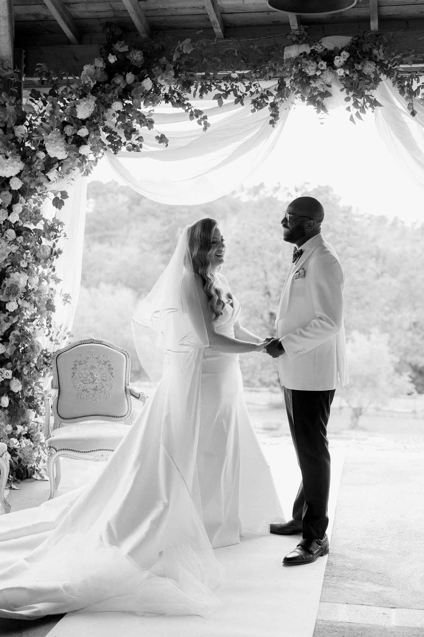 Black and white of couple exchanging vows under wooden pergola with floral installation and draped fabric