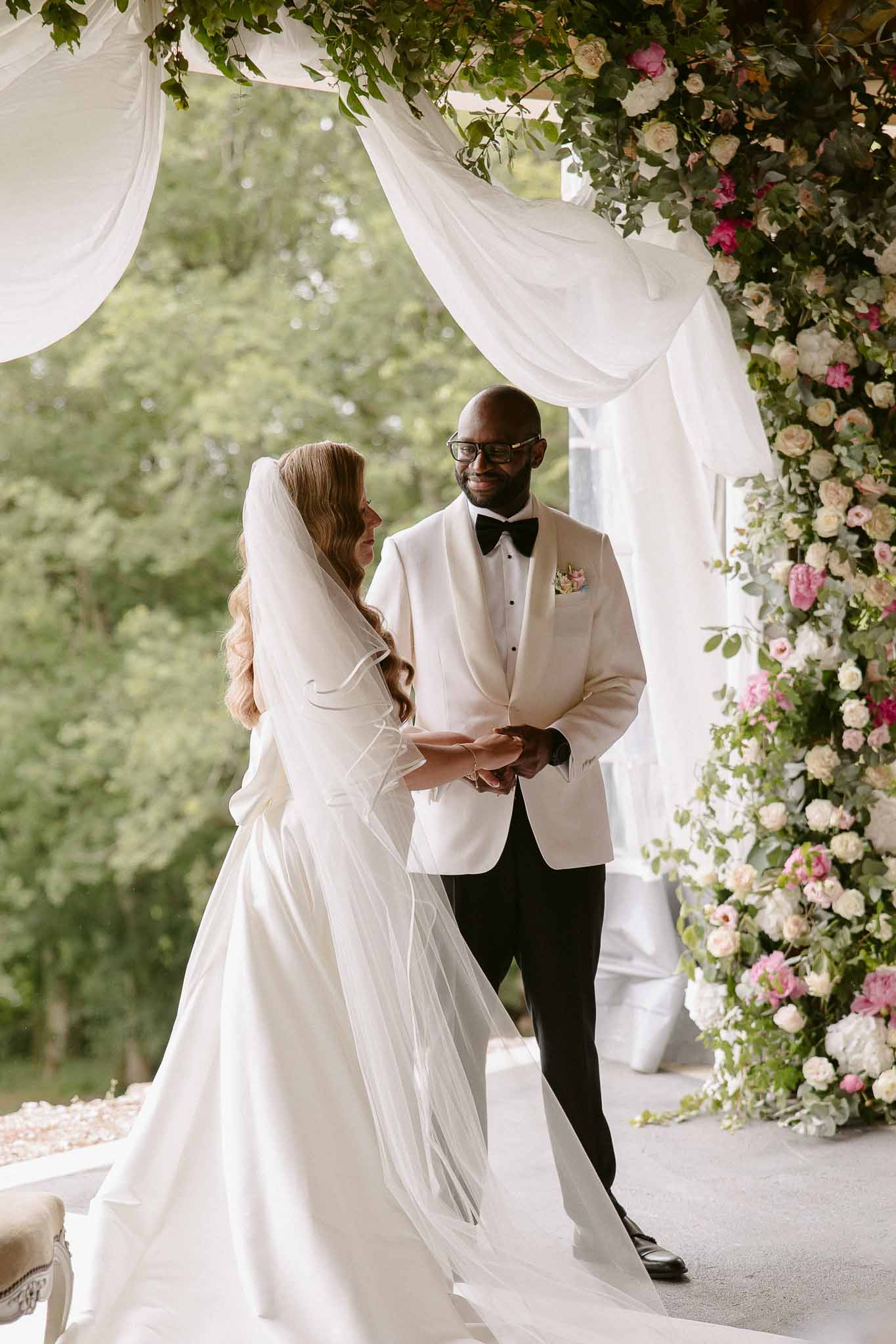 Bride in ivory satin gown and groom in ivory dinner jacket exchange vows under cream and pink rose arch