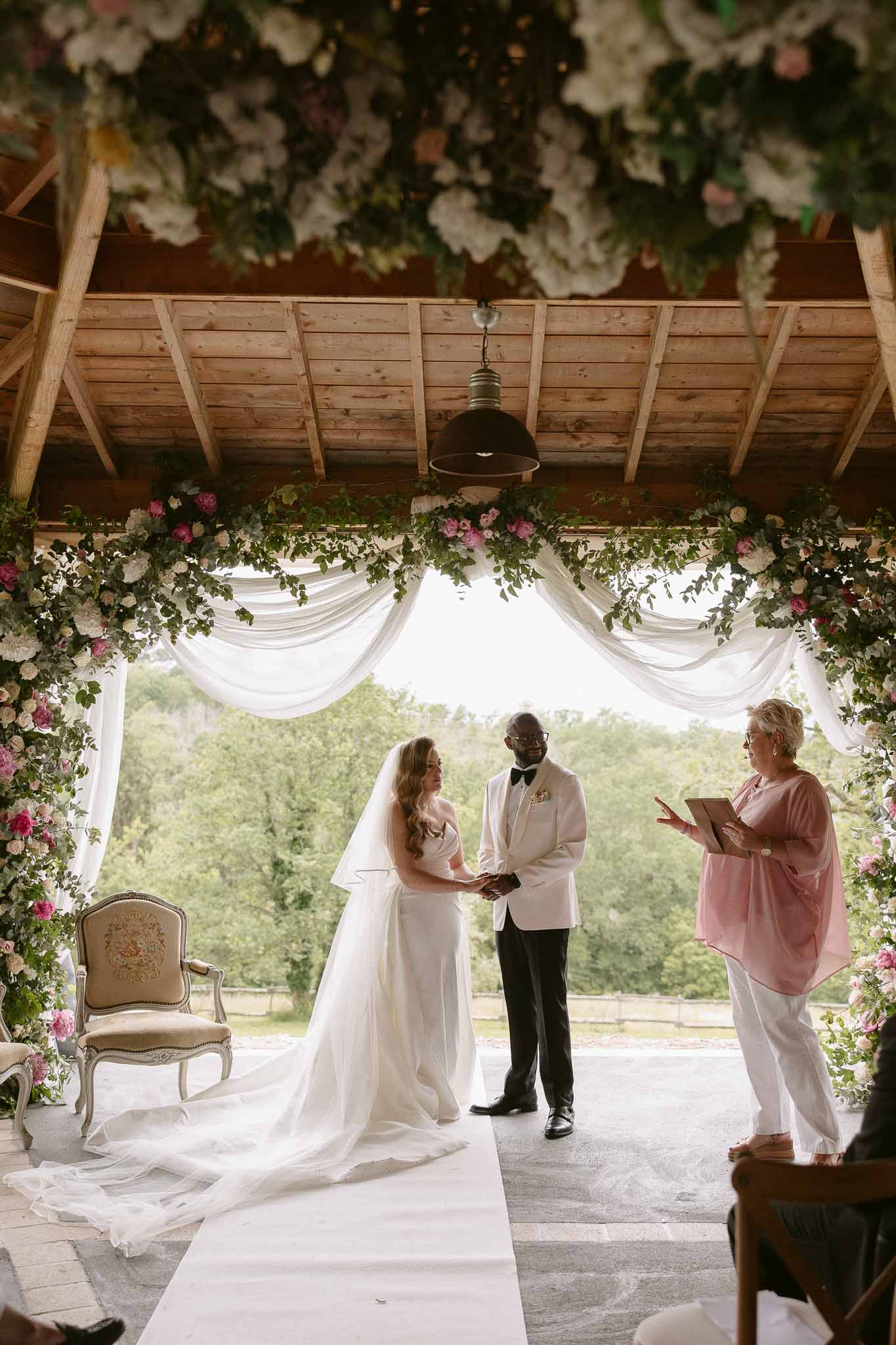Couple at altar under white and pink rose arch with hanging floral installation and white aisle runner
