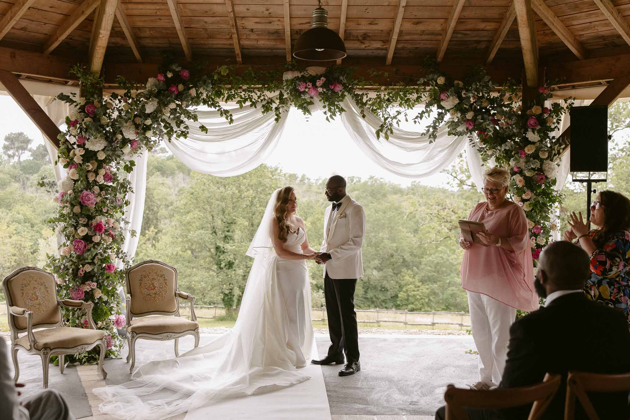 Bride and groom hold hands under wooden pavilion with pink peony and greenery floral arch during ceremony