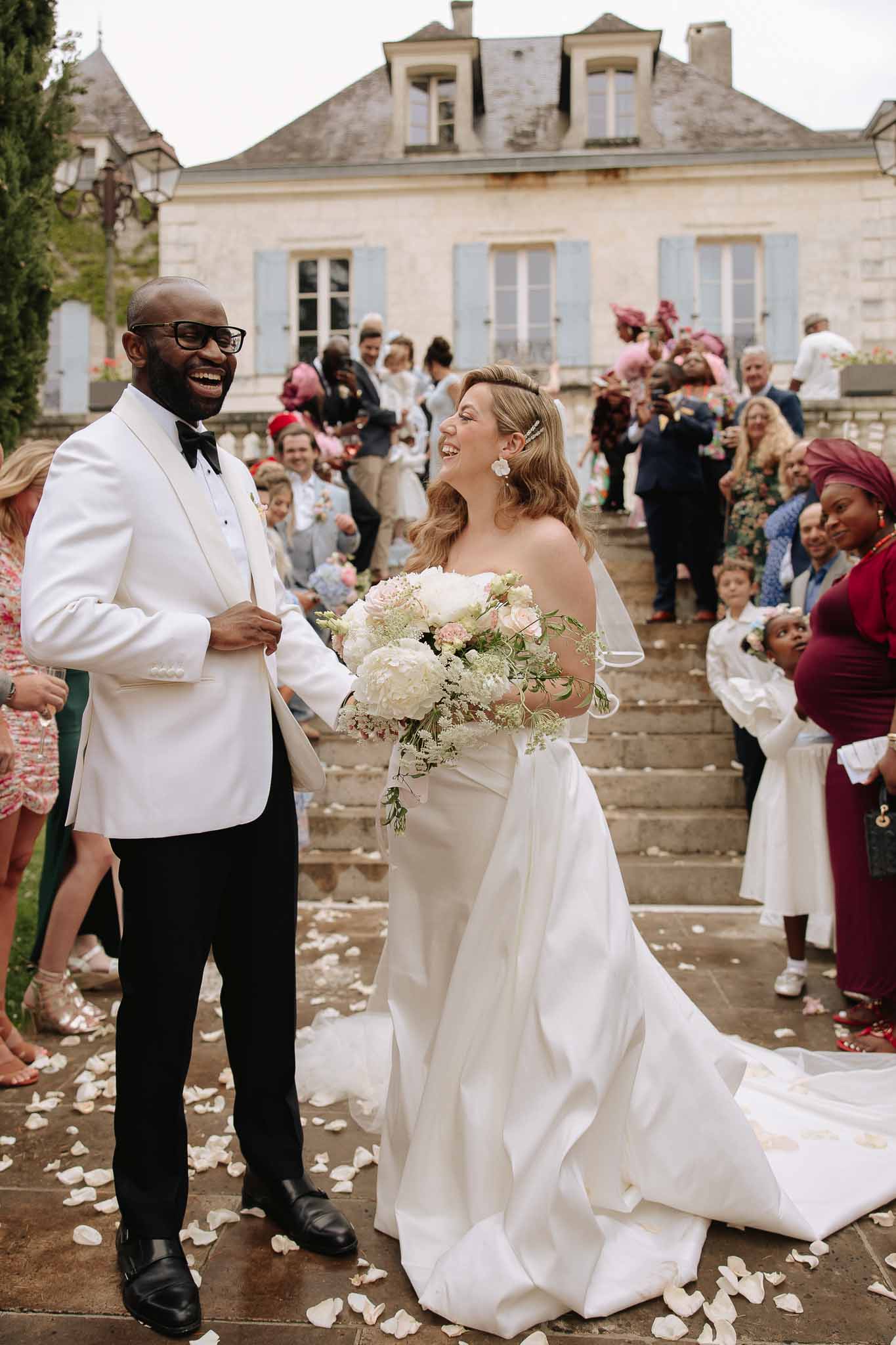 Couple laughing through petal send-off on chateau steps with ivory peony bouquet and blue shutters