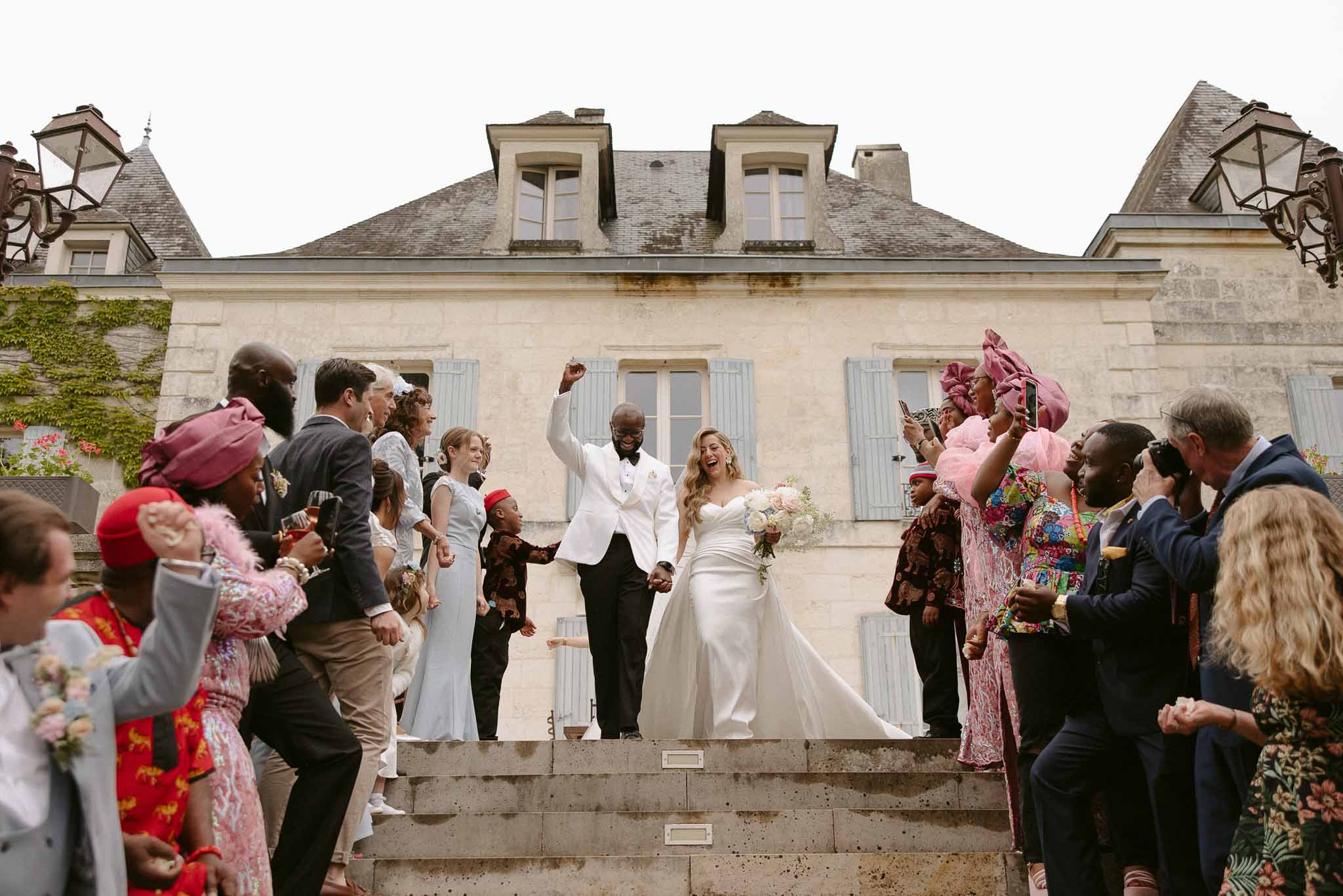 Couple exits down chateau steps with cheering multicultural guests in colorful attire and traditional clothing
