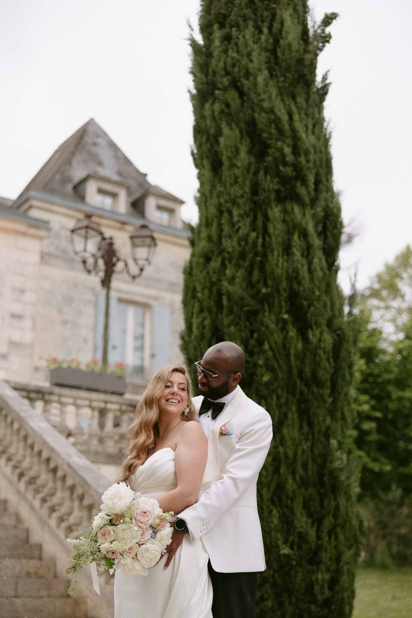 Couple at chateau steps with bride holding ivory peony bouquet and groom in white dinner jacket