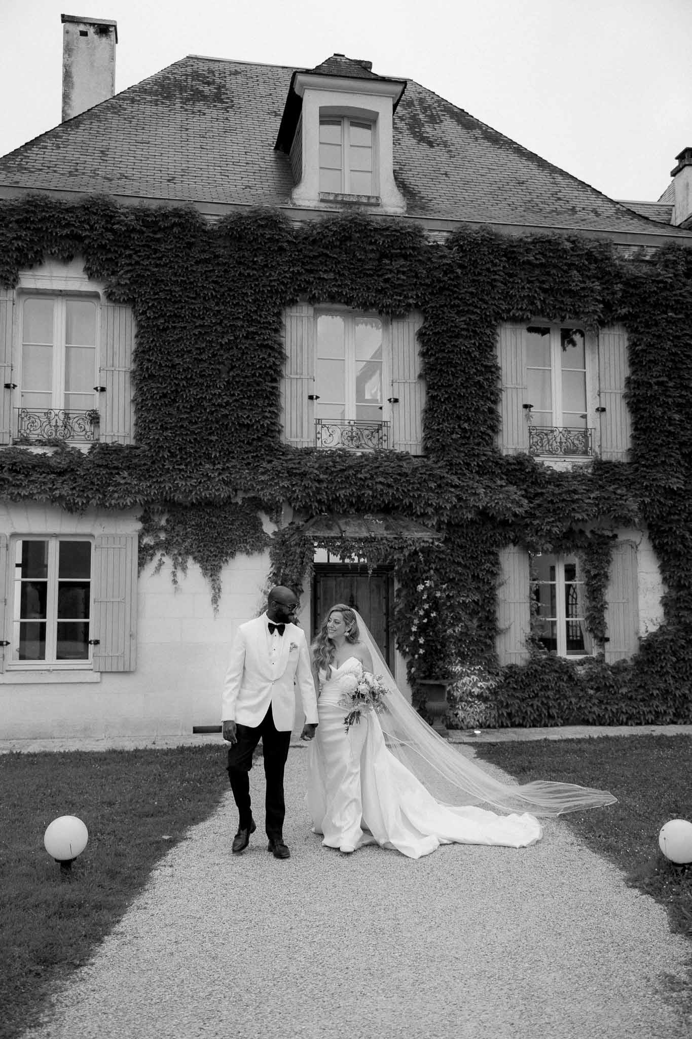 Black and white portrait of couple before ivy-covered manor with cathedral veil trailing on pathway