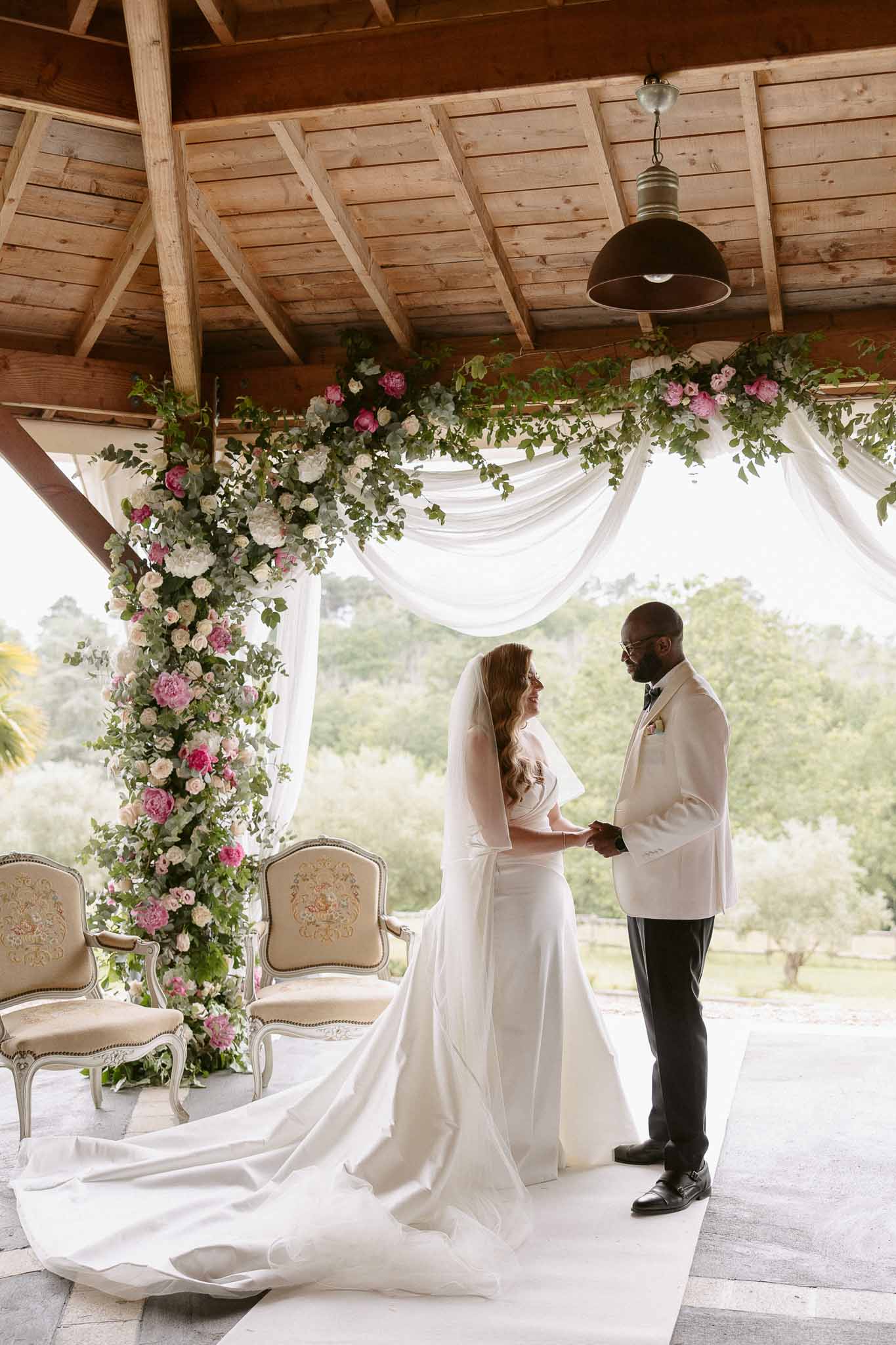 Bride and groom holding hands under rustic pavilion with pink peony and rose floral arch