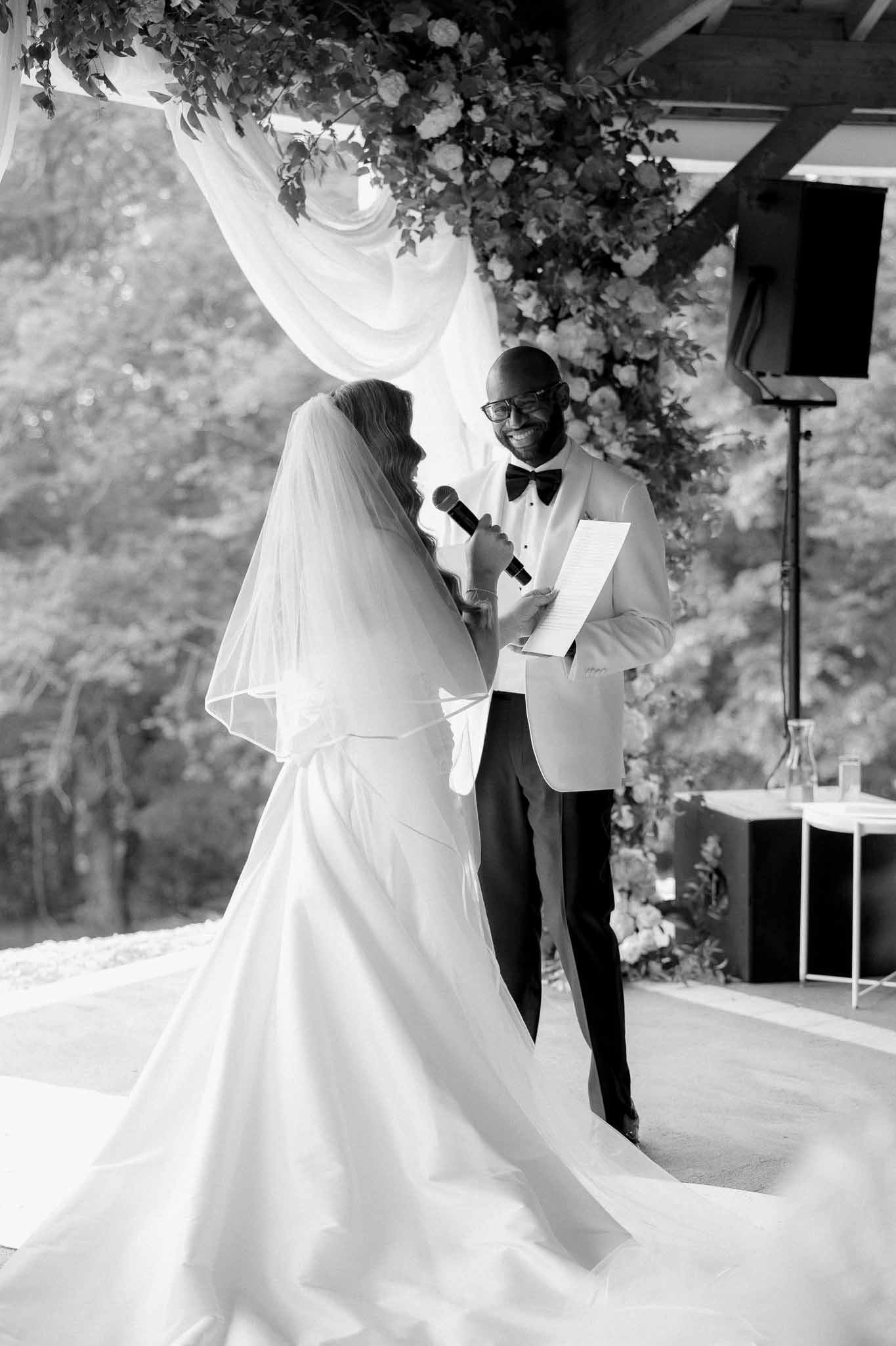 Black and white bride reading vows with microphone beside smiling groom under floral-draped ceremony arch
