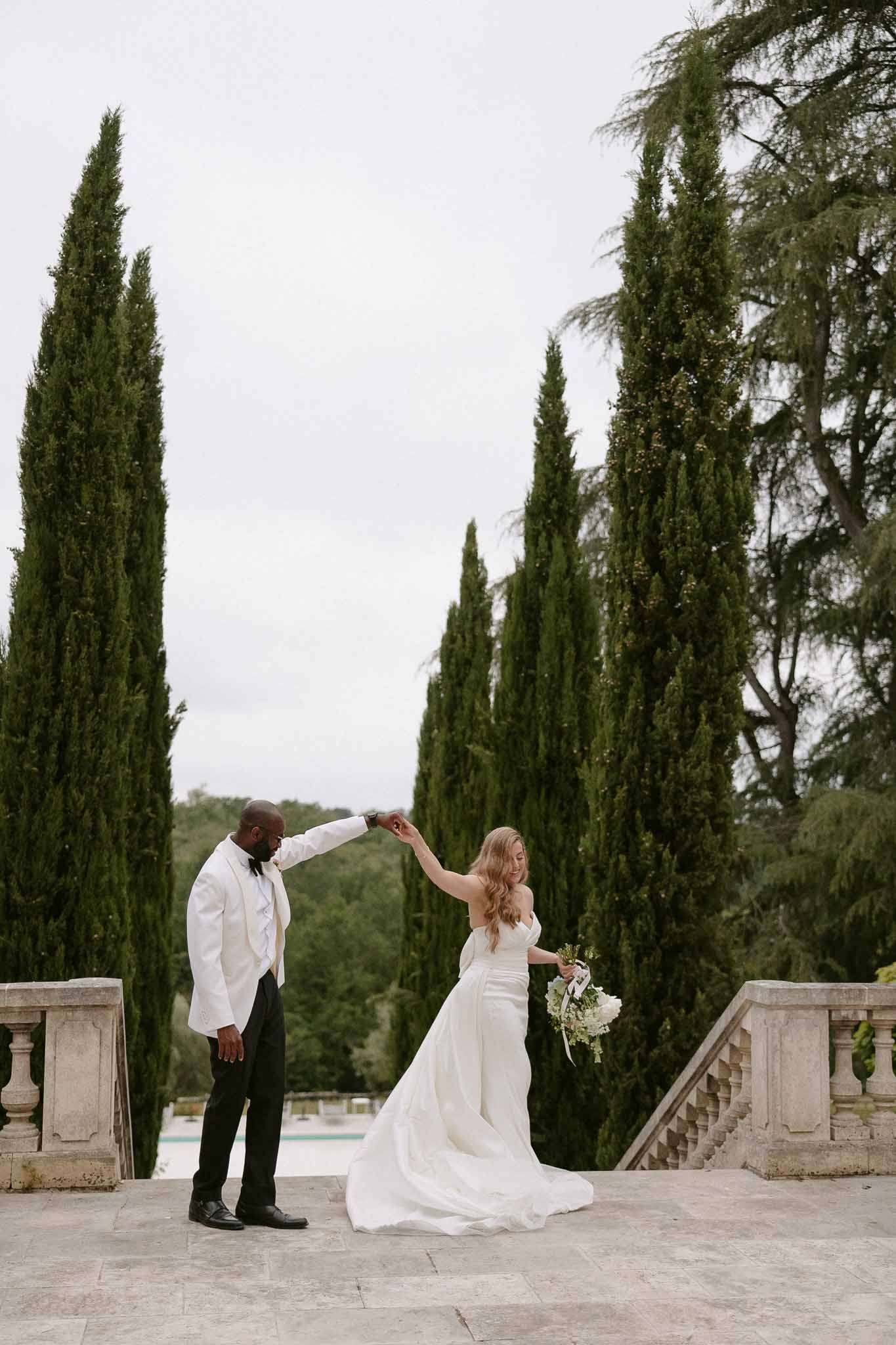 Groom in white dinner jacket spins bride in strapless ivory satin gown on stone terrace with pool and cypress trees