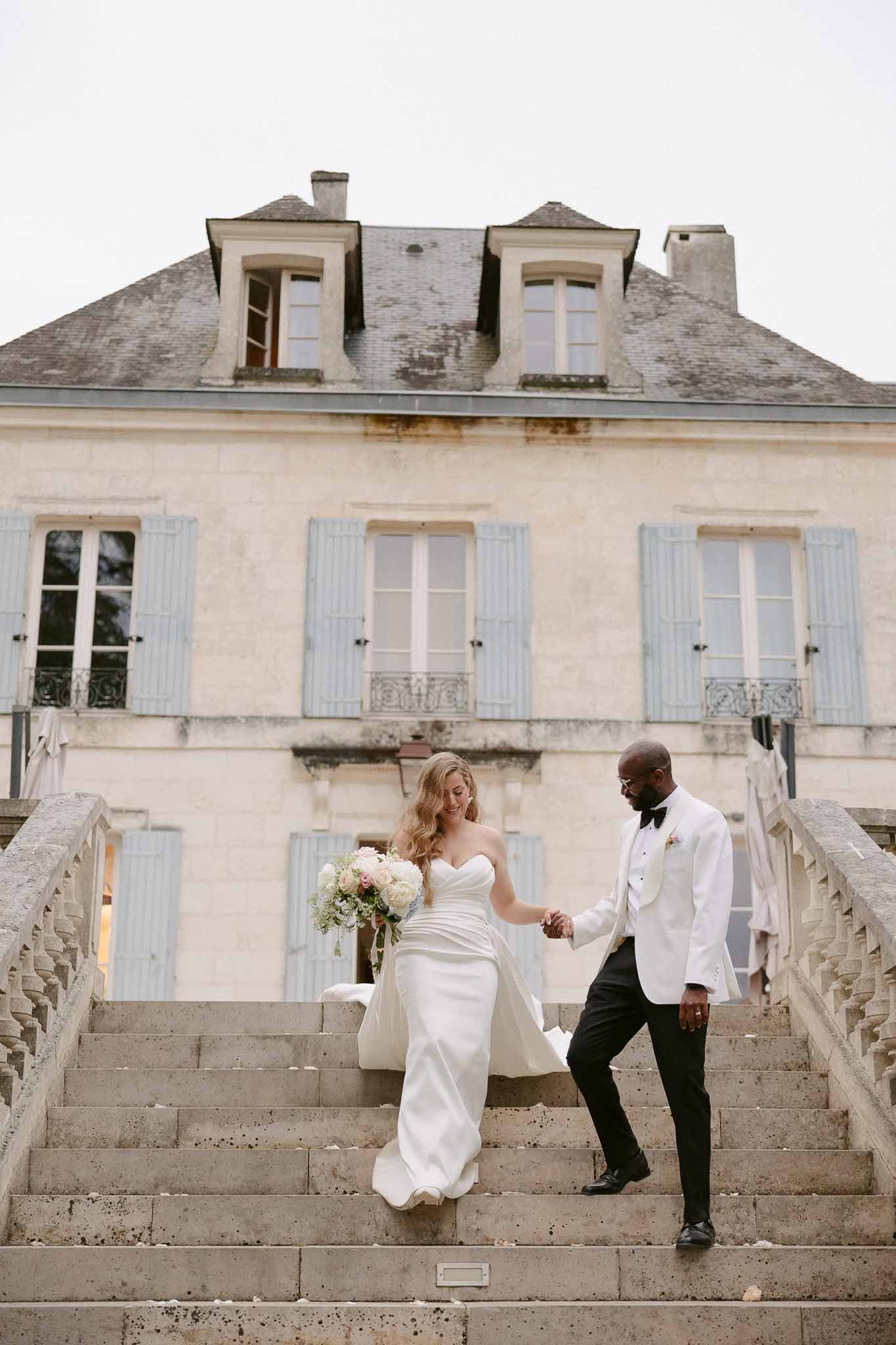 Couple descending chateau stone staircase with bride holding peony and blush rose bouquet and groom carrying train