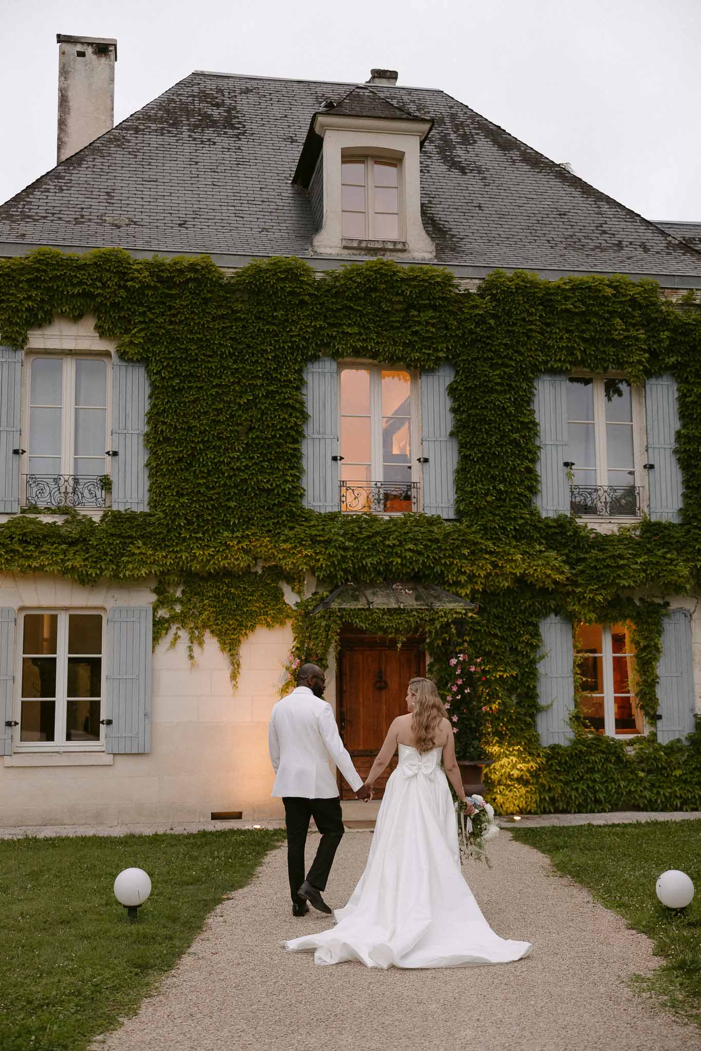 Couple from behind walking toward ivy-covered manor at dusk bride in ballgown with bow detail groom in white dinner jacket