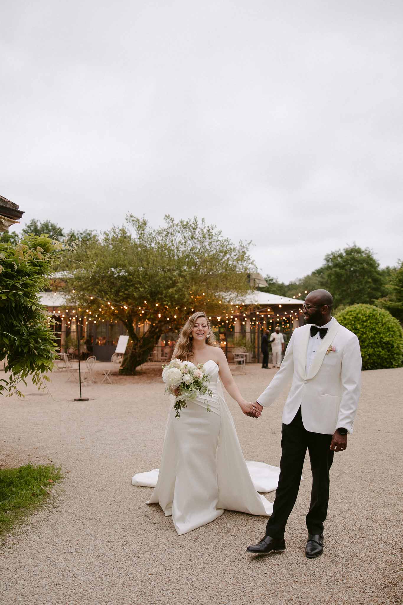 Couple walking on gravel, groom in white tuxedo jacket, bride with hydrangea and blush rose bouquet