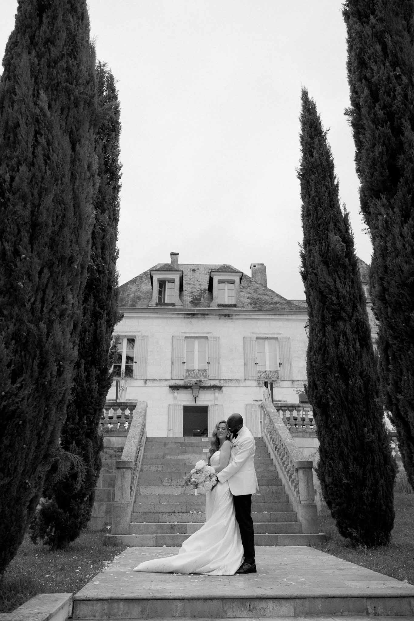 Black and white couple portrait at base of chateau staircase framed by cypress trees