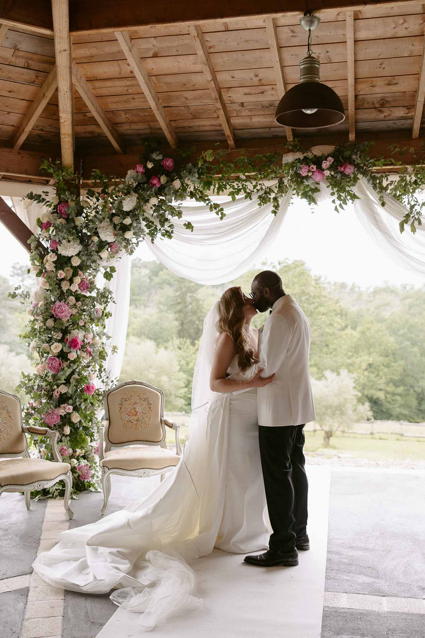 Bride and groom first kiss under hot pink peony and blush rose arch in covered wooden pavilion