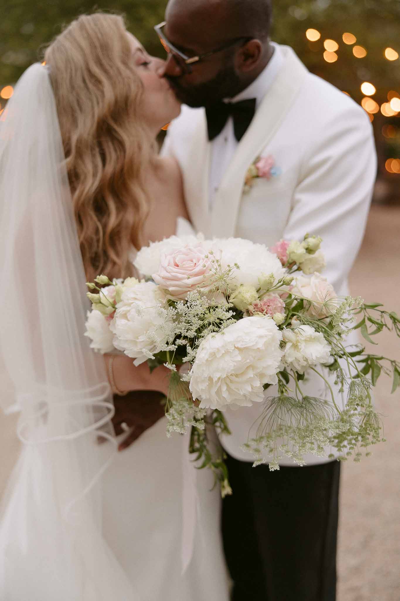 Couple kisses with peony and garden rose bouquet sharp in foreground and string lights bokeh behind