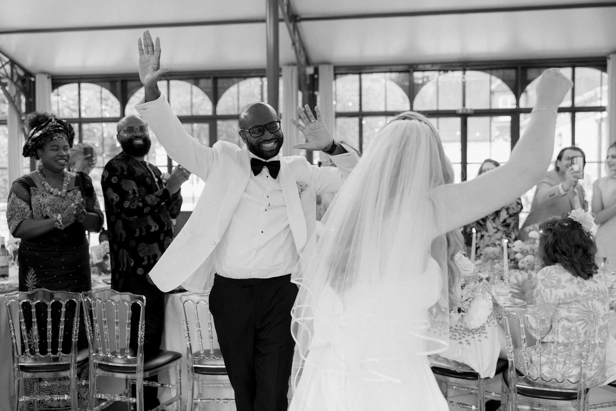 Black and white image of couple making grand entrance to reception with arms raised and guests cheering