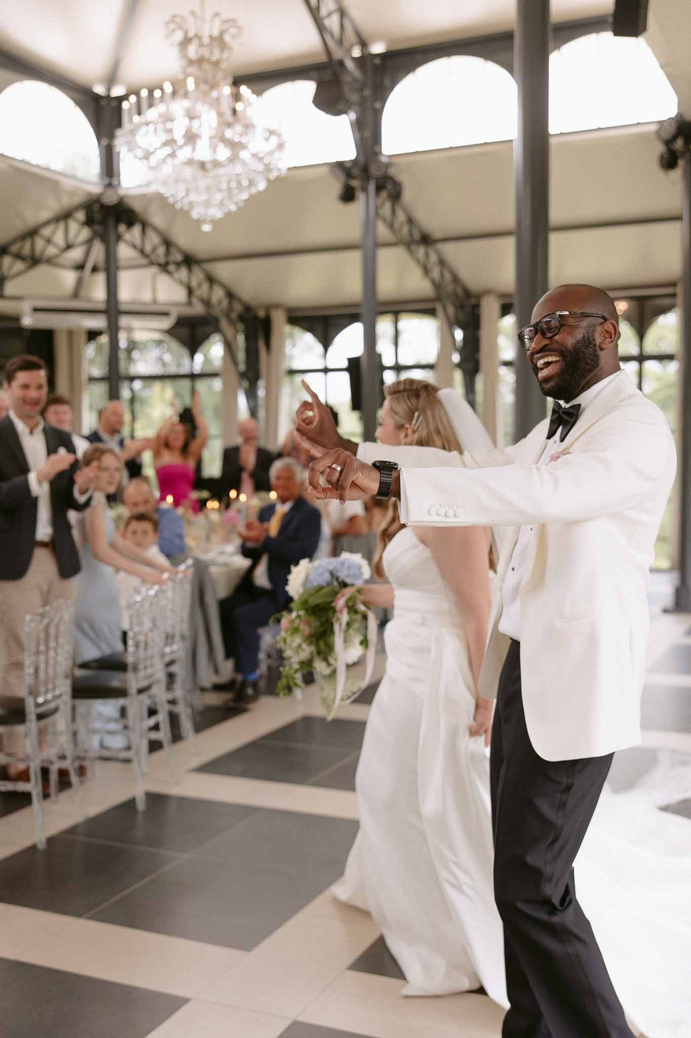 Bride and groom enter glass-and-iron pavilion reception as guests cheer at round tables with ghost chairs
