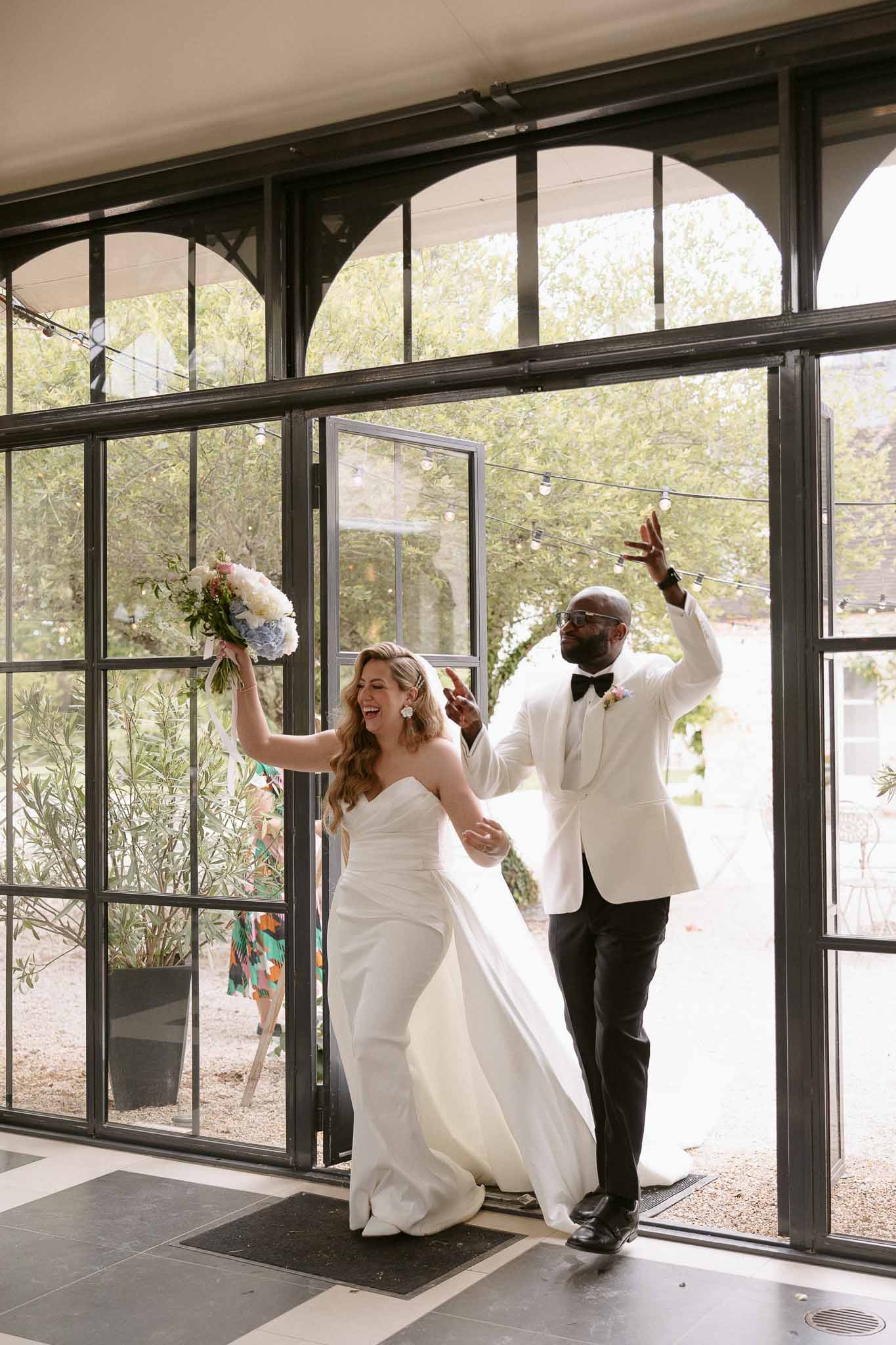 Couple laughing with arms raised entering reception through arched glass doors holding blue hydrangea bouquet