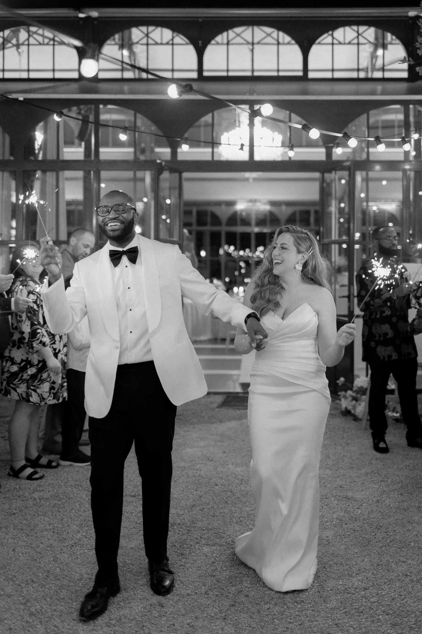 Sparkler send-off with couple walking through guest lines at night outside glass conservatory in black and white