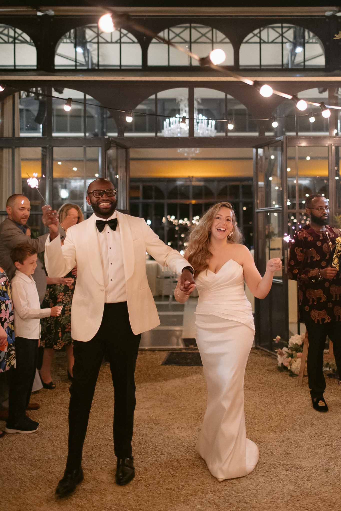 Bride and groom make grand entrance through sparkler-lit aisle at indoor reception with arched windows