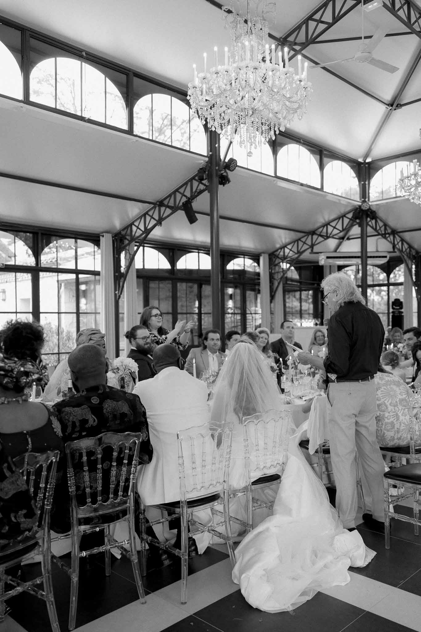 Black and white speech moment in conservatory with crystal chandelier, ghost chairs, and candlelit banquet table