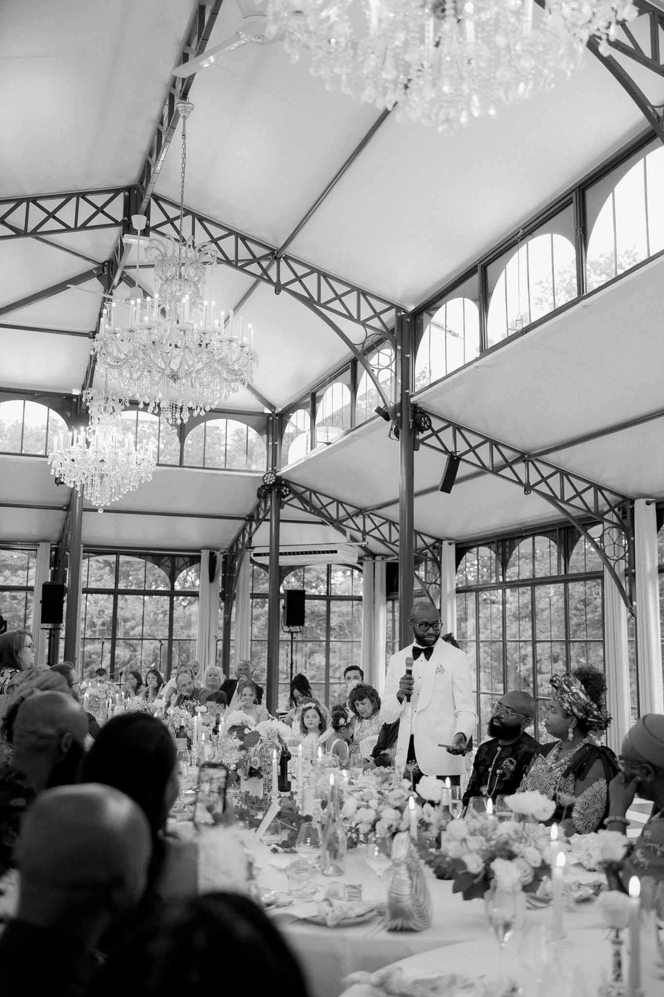 Black and white speech moment in orangerie venue with crystal chandeliers and seated guests