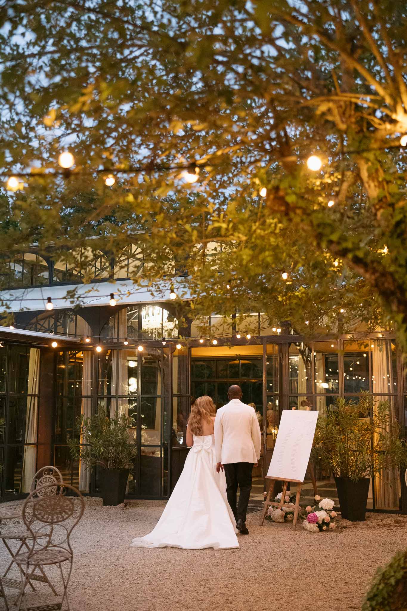 Couple walking toward glass greenhouse reception venue at dusk with festoon lights and seating chart