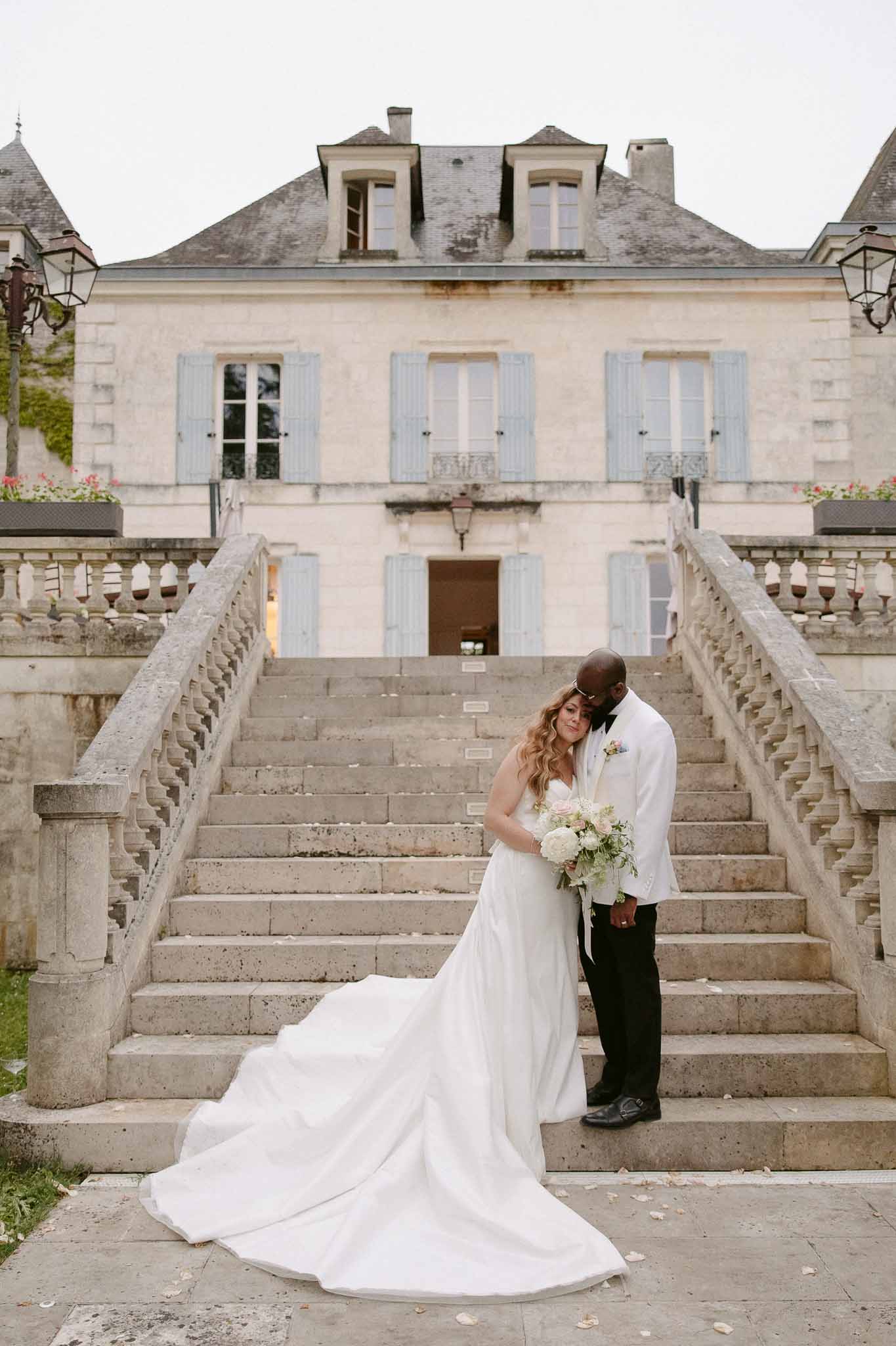 Bride with hydrangea bouquet and groom in white dinner jacket on chateau stone staircase with scattered petals