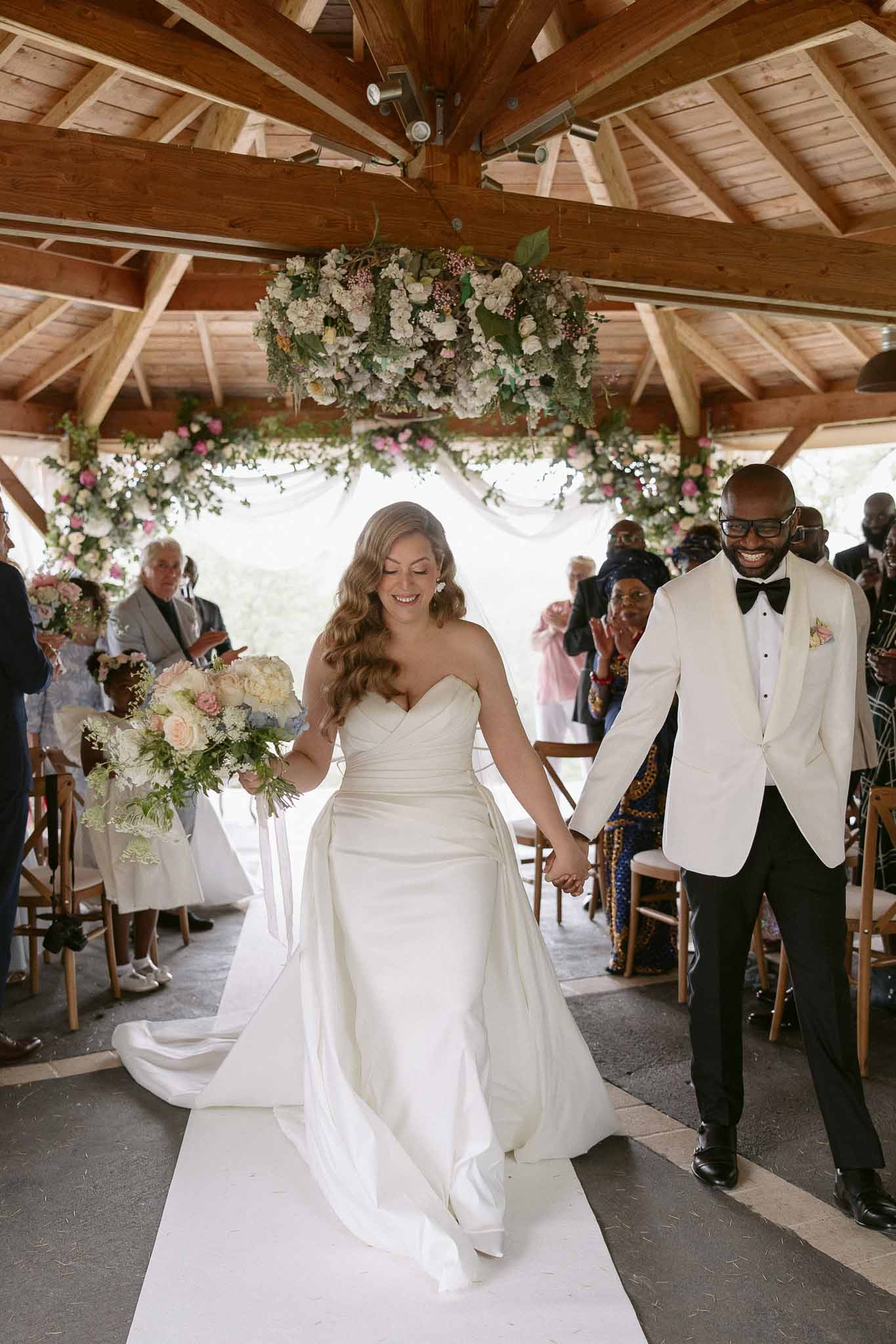 Couple recessioning under floral chandelier and fabric arch bride in ruched satin gown with cream rose bouquet guests appl...
