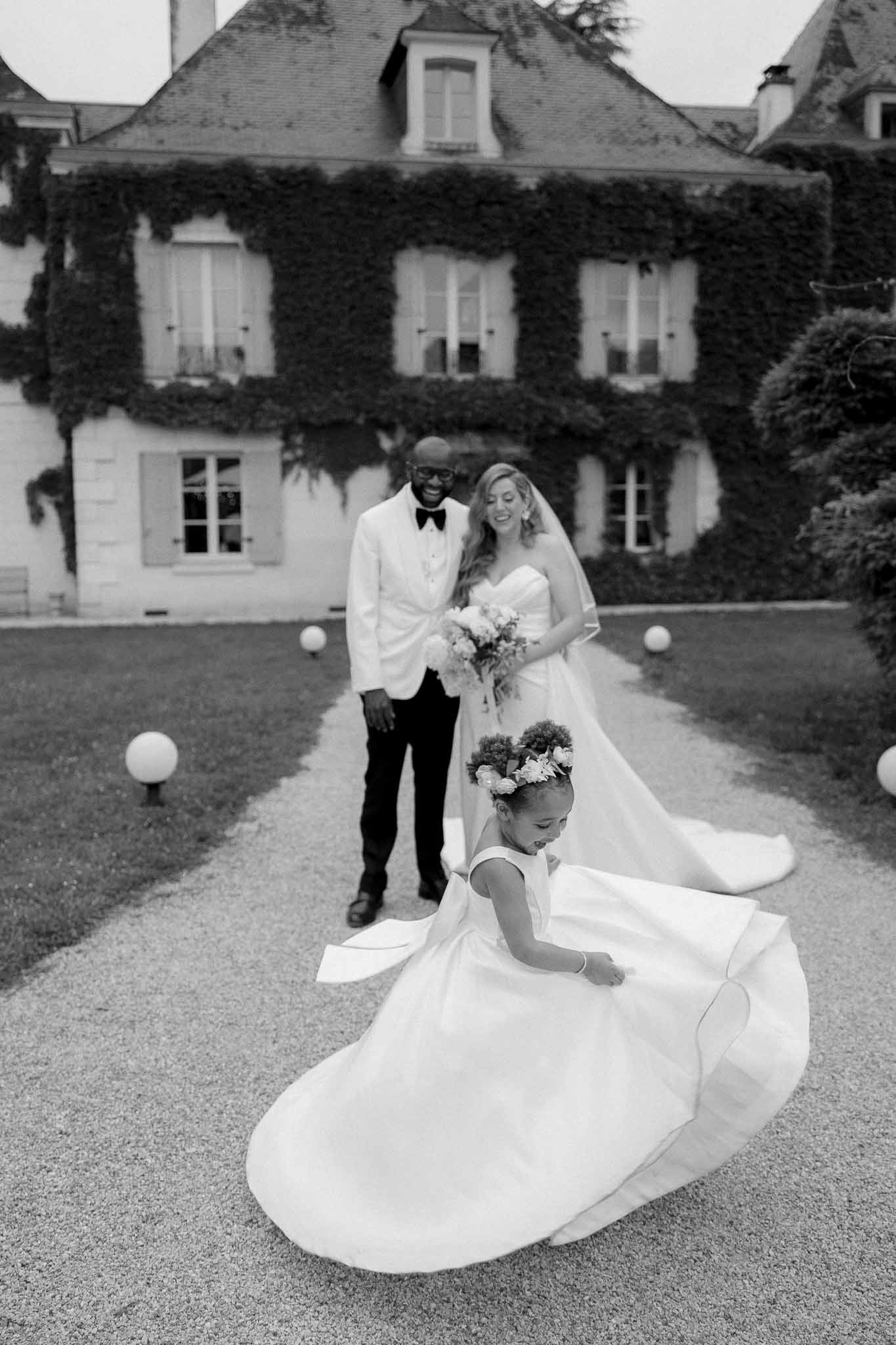 Black and white shot of flower girl adjusting bride's train as couple smiles on chateau grounds