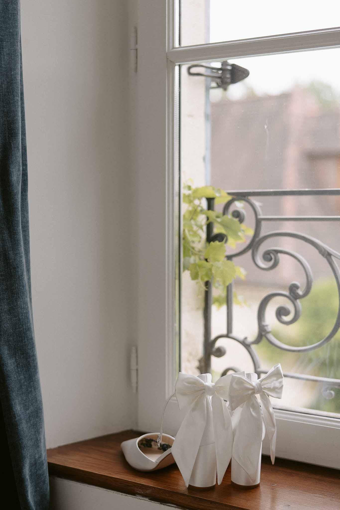 White bridal bow mules and ankle-strap heels on a wooden windowsill with wrought-iron scrollwork outside