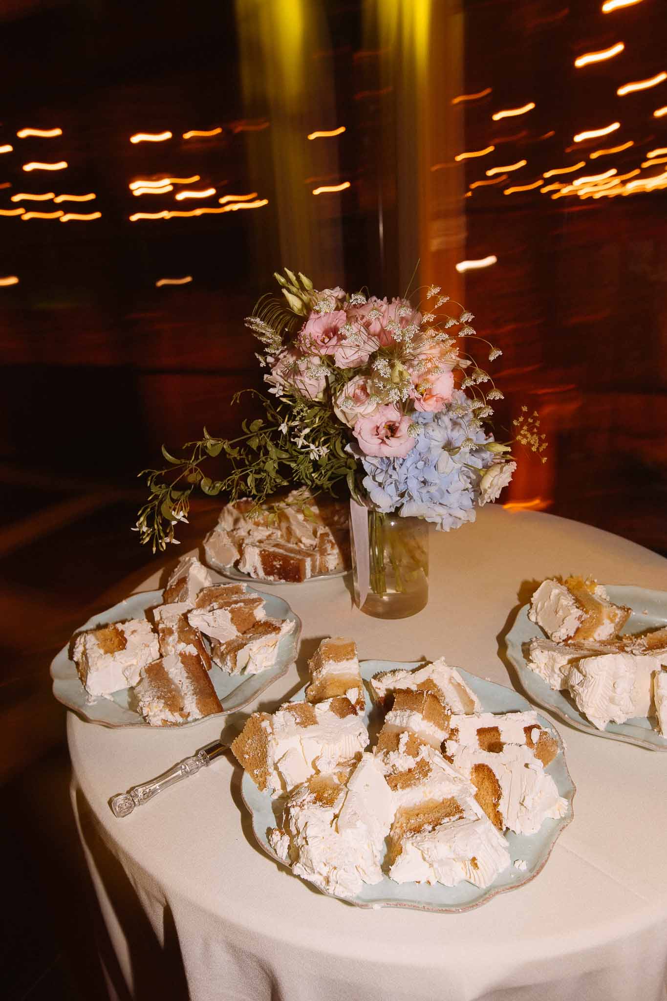 Dessert table with sliced layered cake on blue plates, blush and blue floral arrangement, festoon lights behind