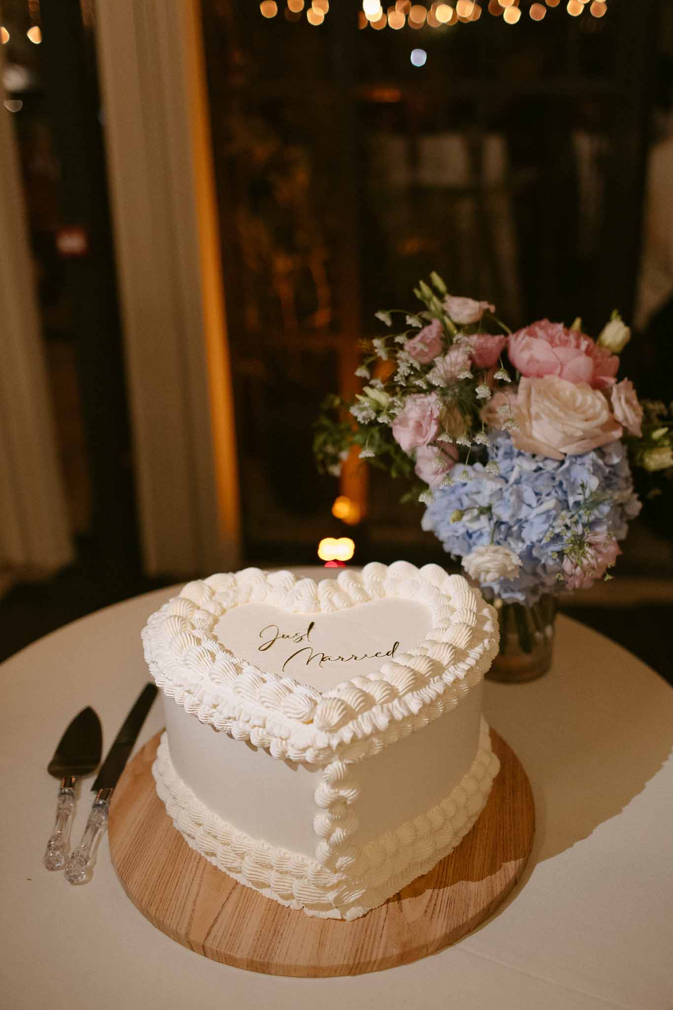 Heart-shaped white wedding cake with gold Just Married script beside blue and pink bridal bouquet