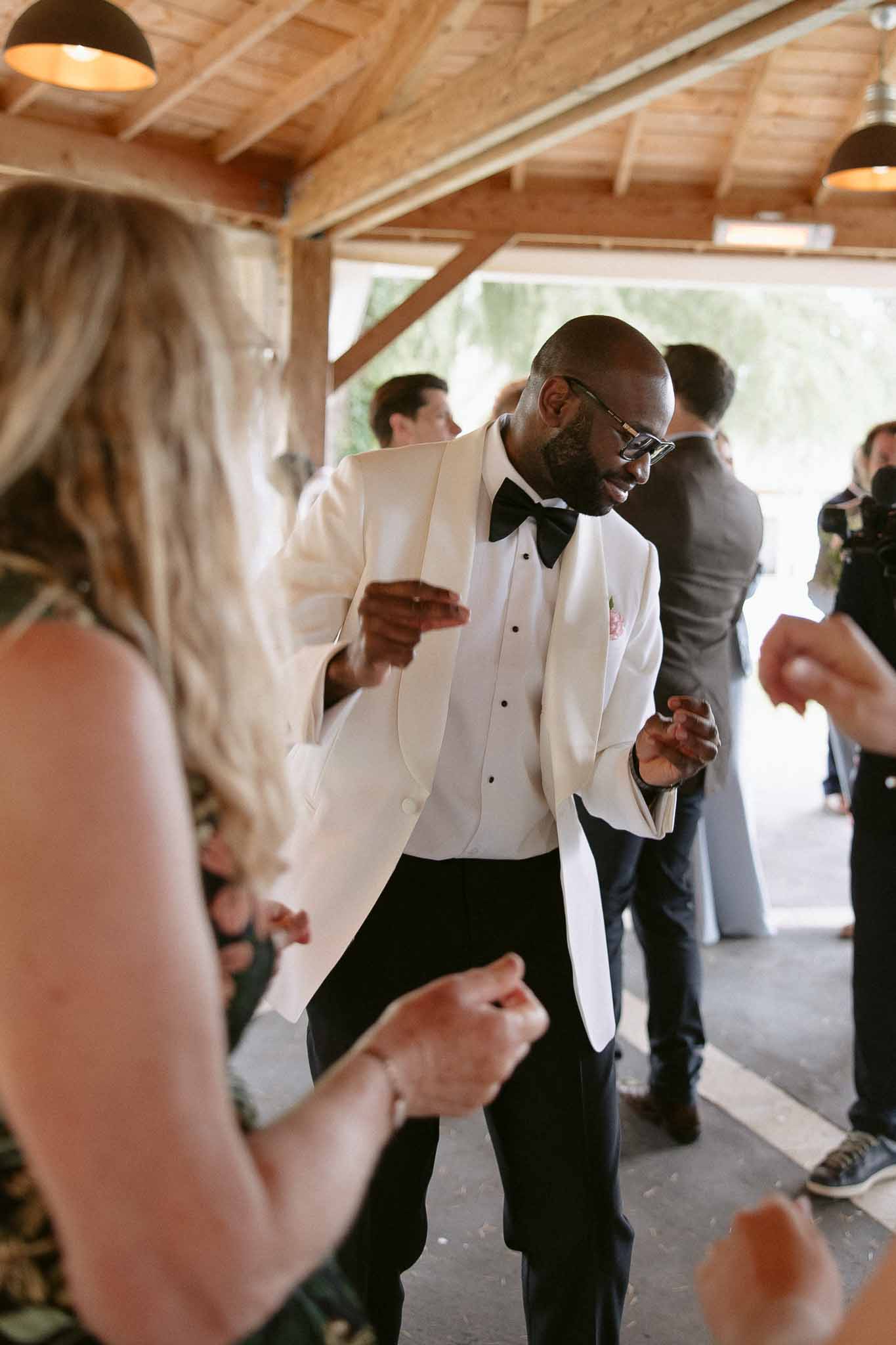 Man in cream dinner jacket dancing energetically at barn-style reception venue with exposed wooden beams