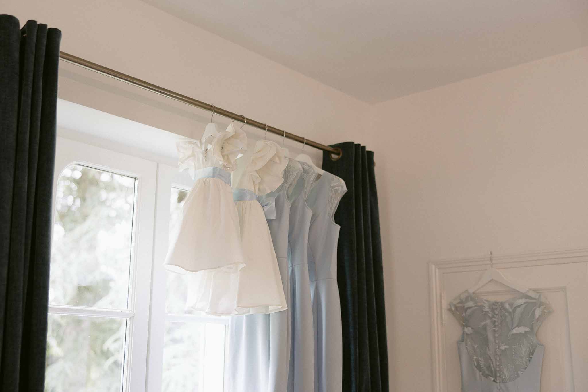 Powder blue bridesmaid dresses and white flower girl dress hanging on curtain rod by window