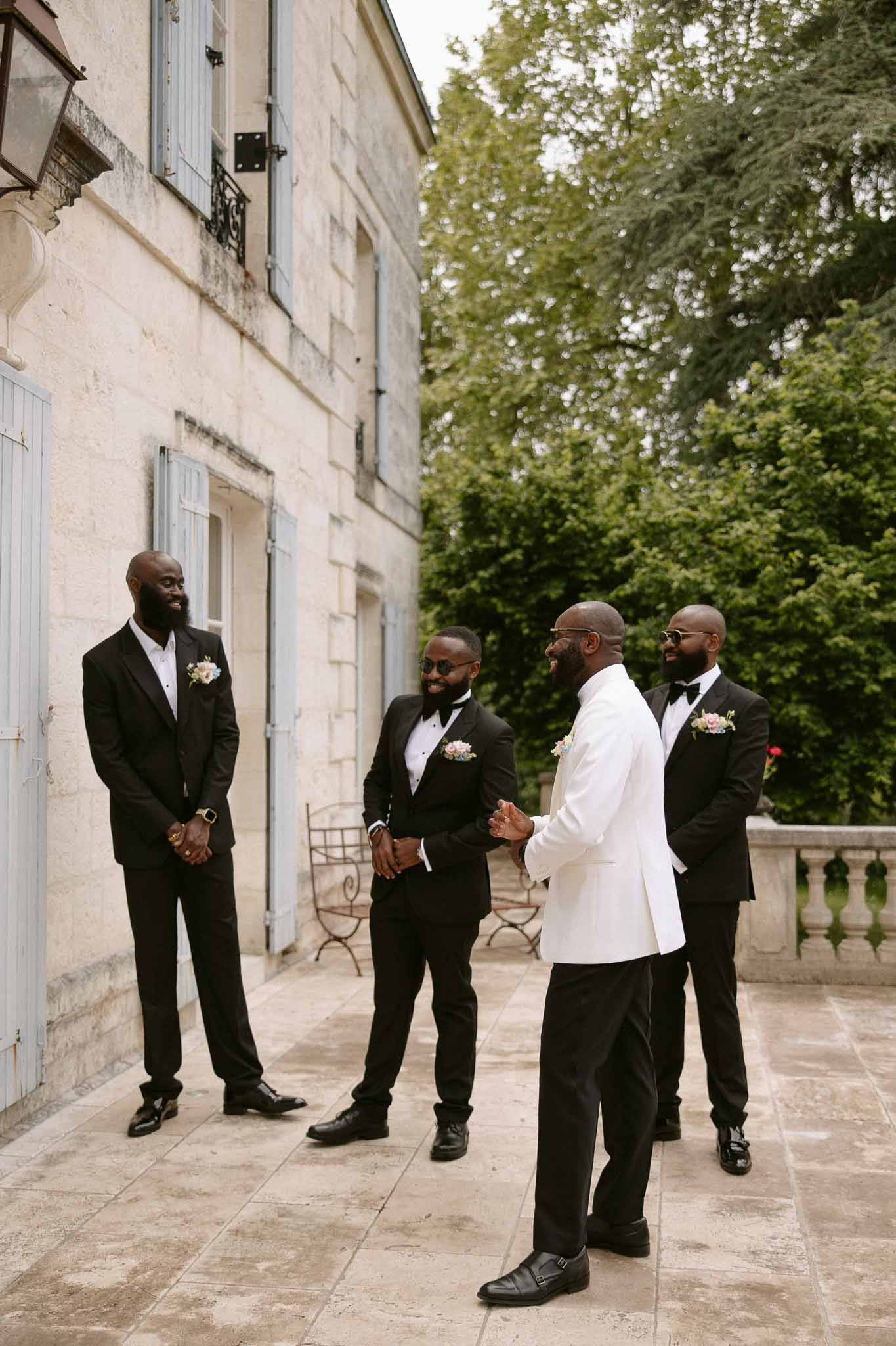 Groom in white dinner jacket with three groomsmen in black tuxedos laughing on chateau terrace