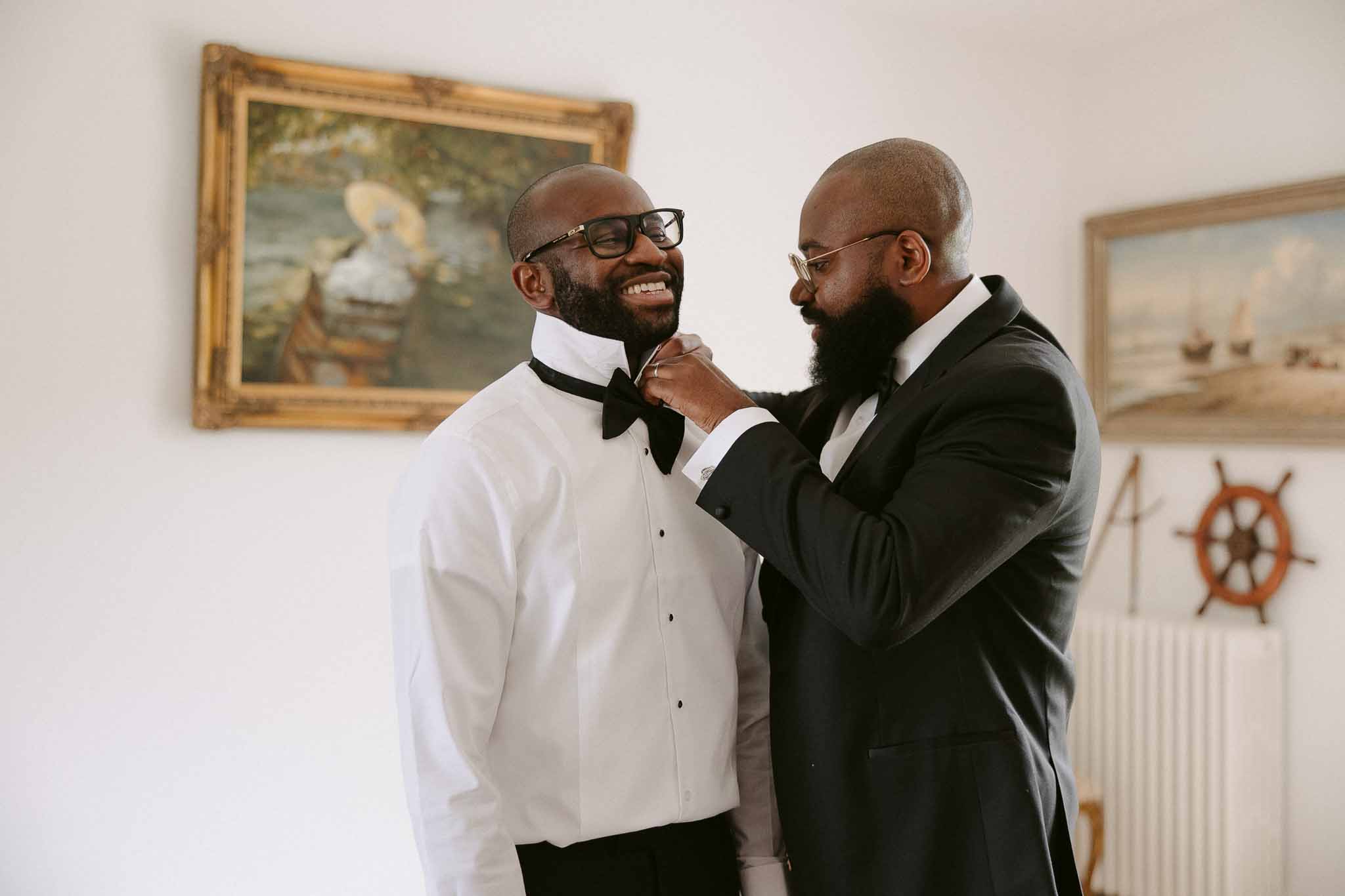 Groomsman adjusts groom's black bow tie while both laugh during getting ready