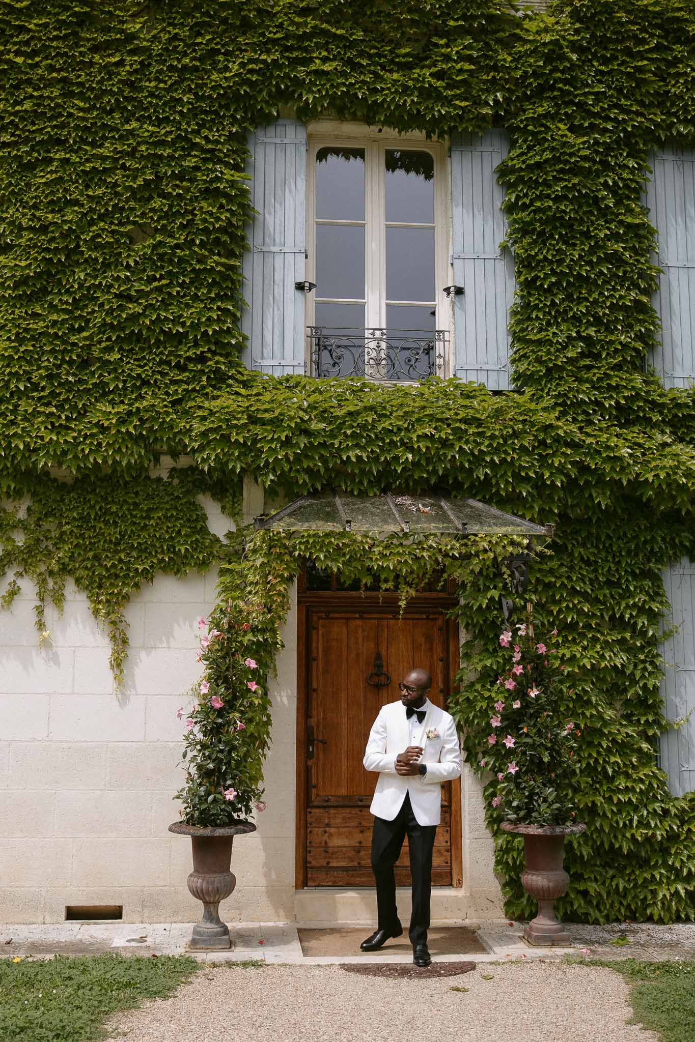 Groom in white dinner jacket reading notes in front of ivy-covered chateau entrance with stone urns