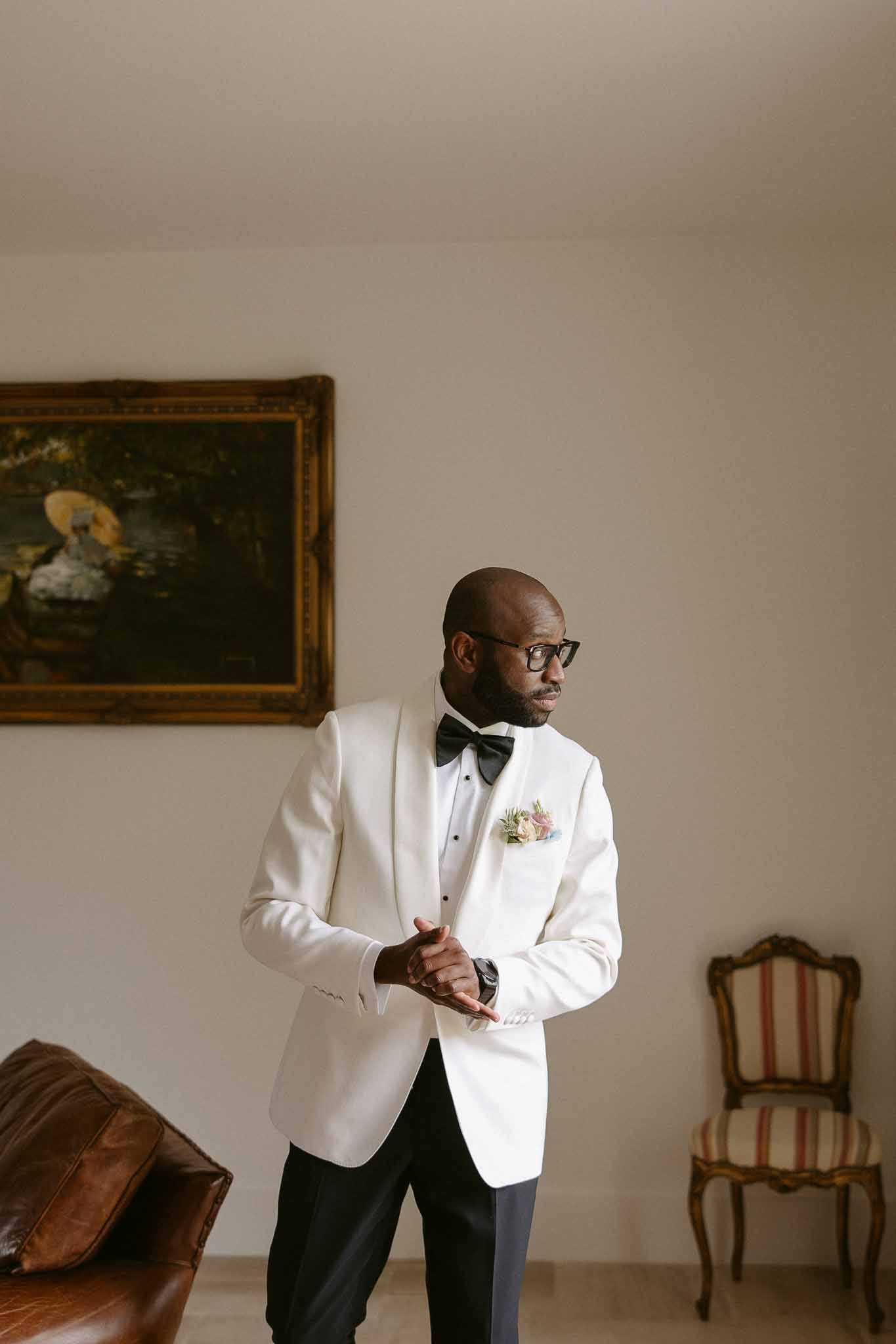 Groom in ivory dinner jacket with shawl lapel and blush boutonniere in classic chateau room with oil painting