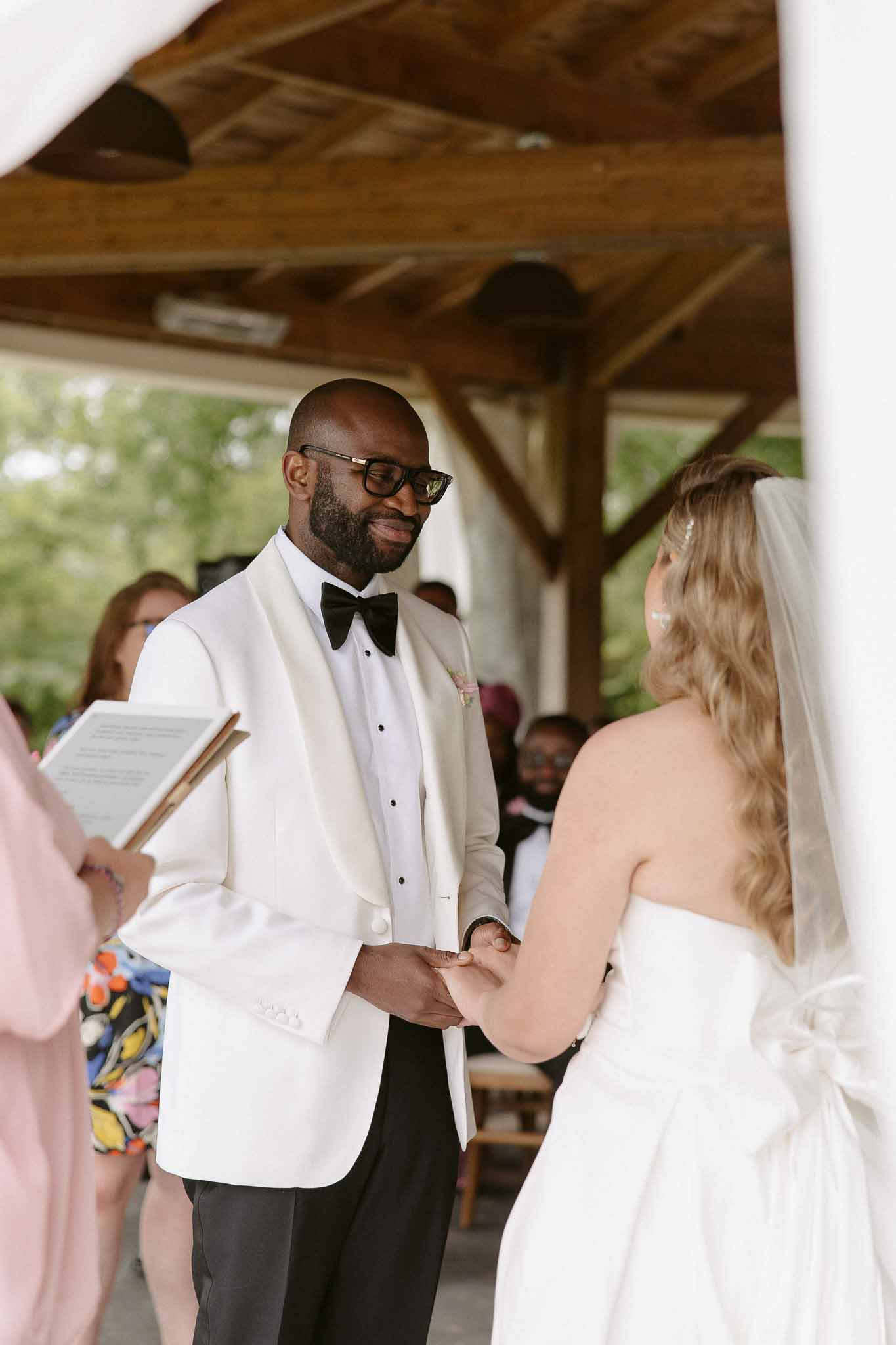 Groom in ivory dinner jacket holding bride's hands and smiling during vows under wooden pavilion
