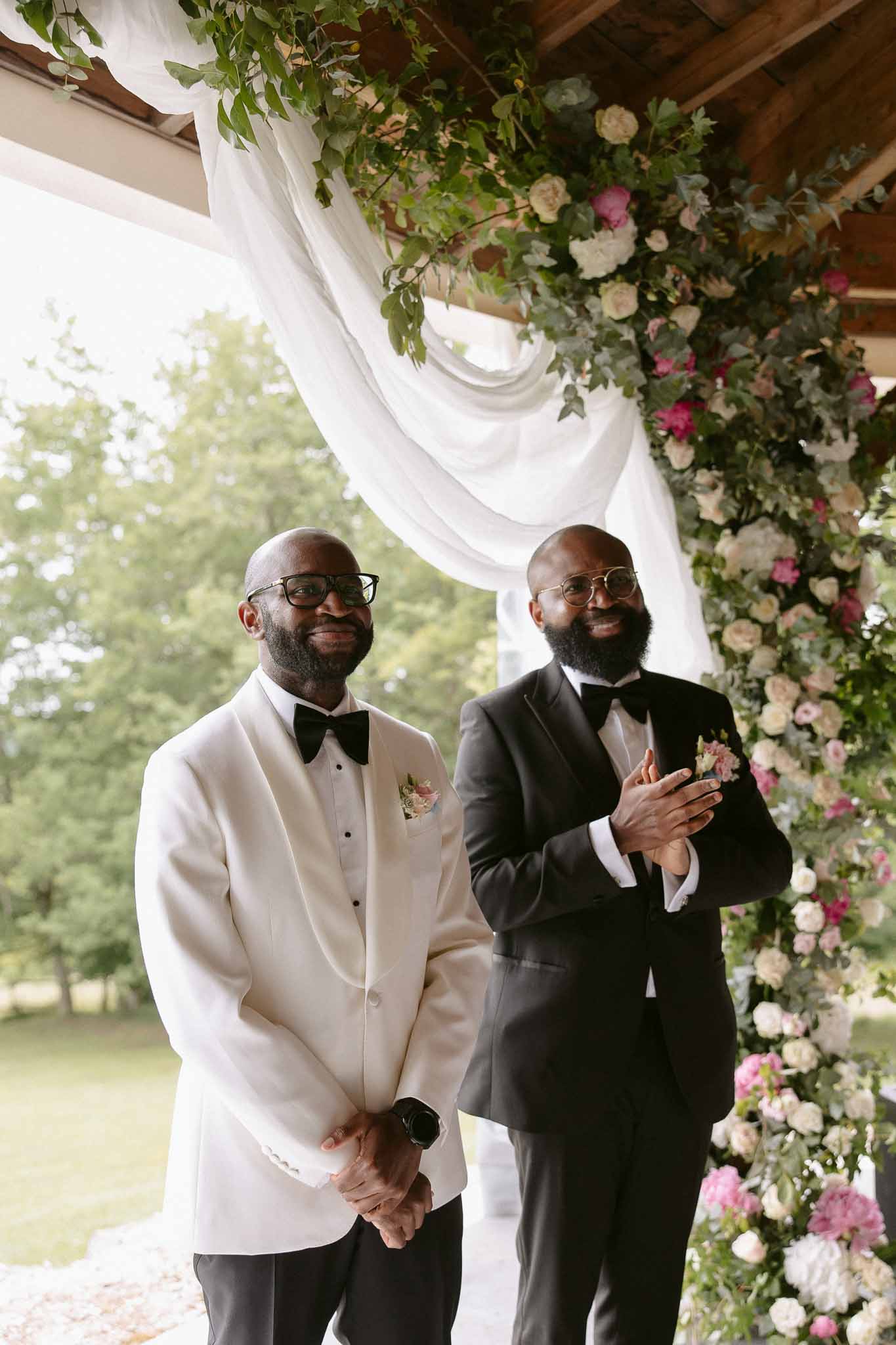 Two grooms smiling at altar beside hot pink rose and ivory peony arch with white fabric draping