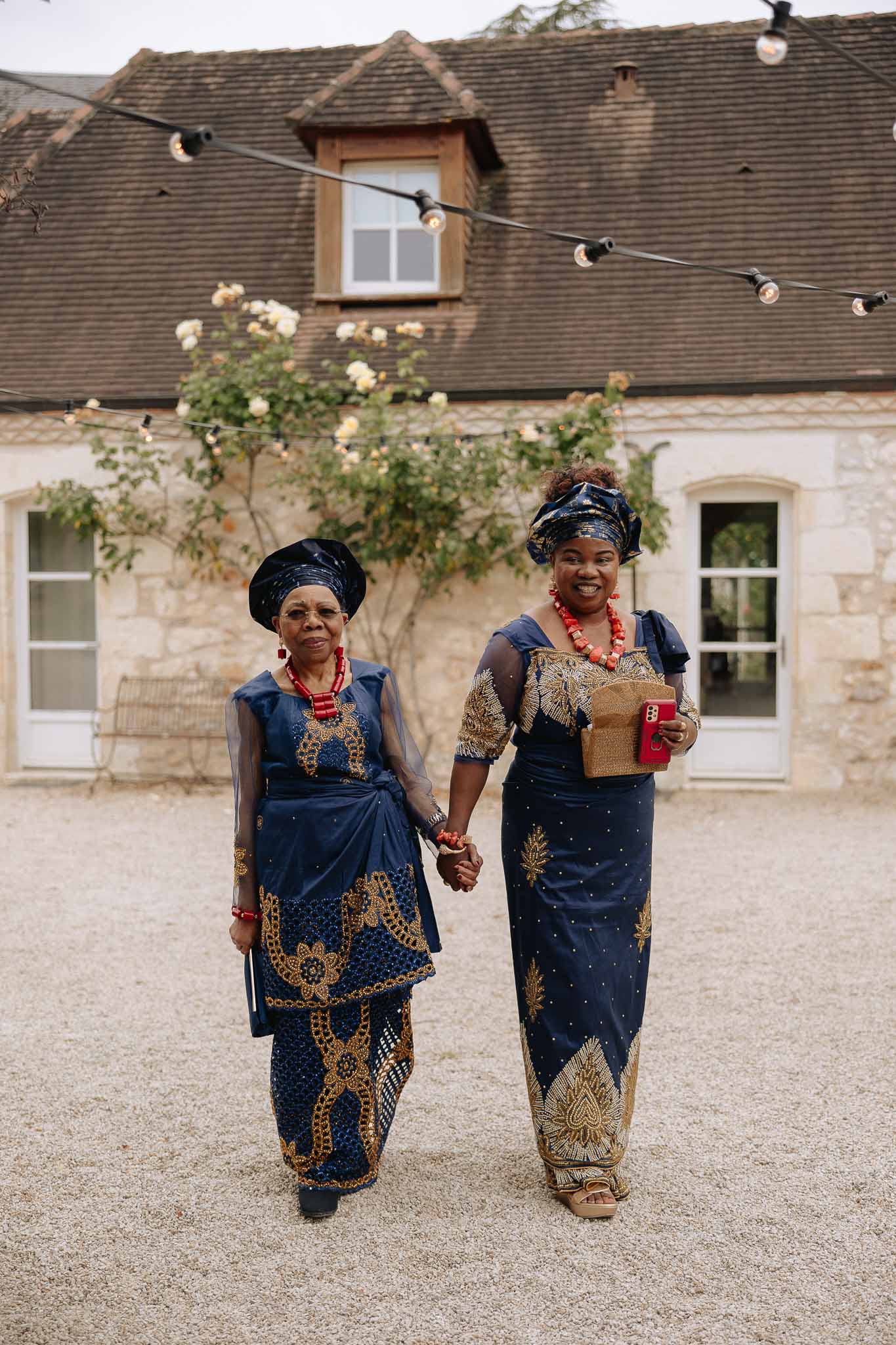 Two guests in navy and gold West African lace with gele head wraps and coral beads walking across stone courtyard